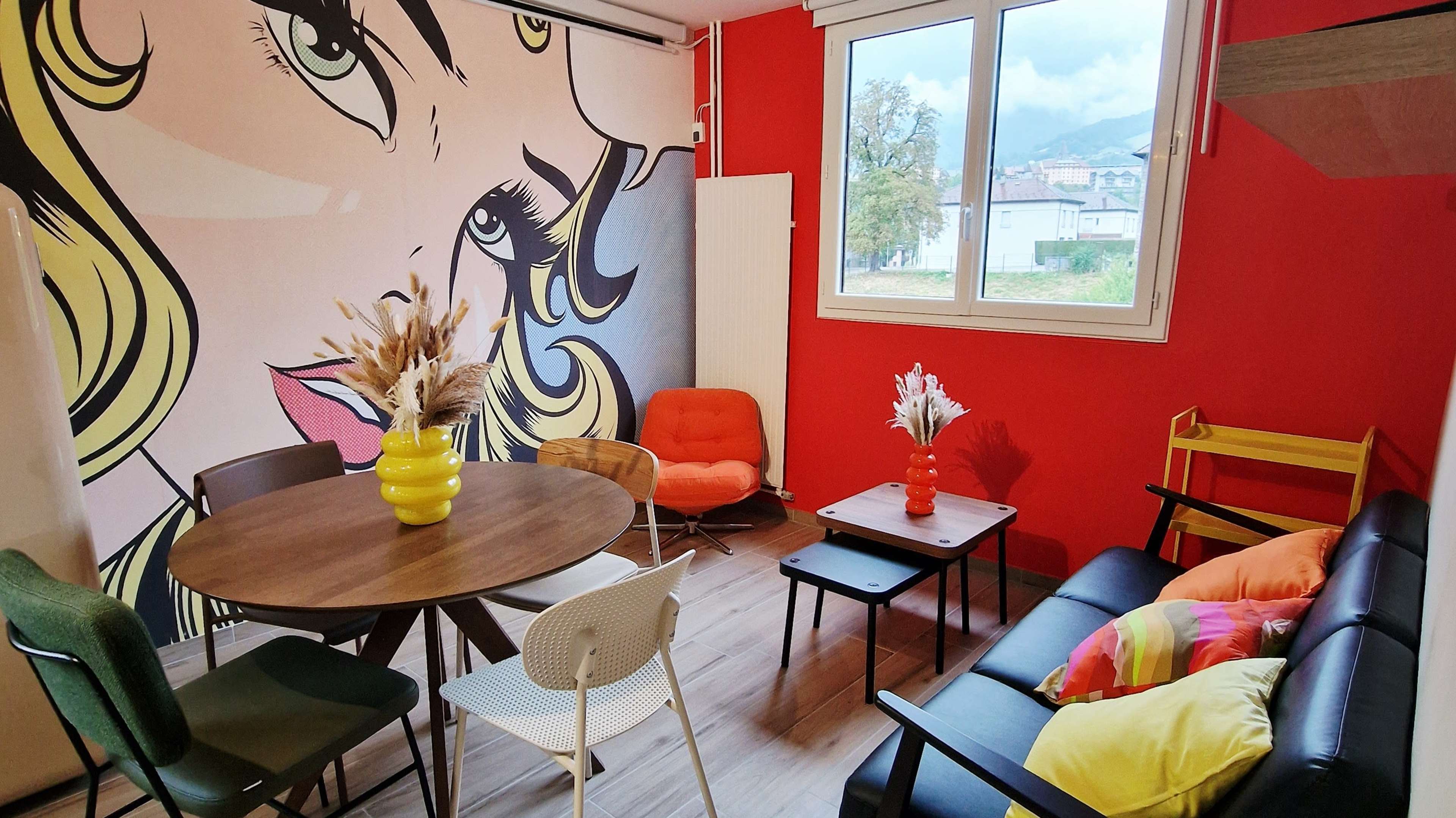 The image shows a colorful indoor seating area featuring a round table surrounded by various chairs, a wall mural of a woman's face, and large windows letting in natural light.