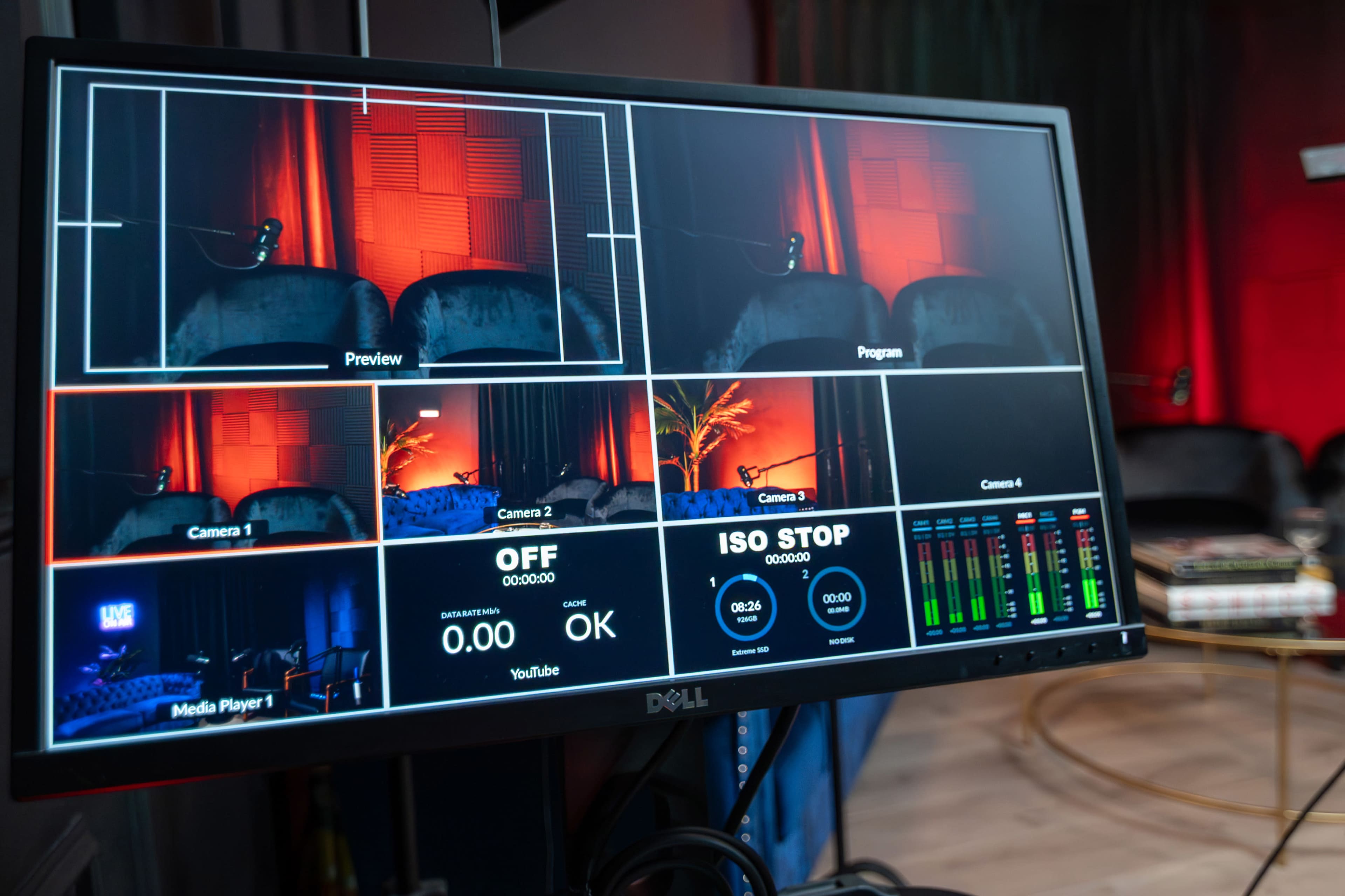 The image shows a computer monitor displaying multiple camera feeds and a media player interface in a studio setting.