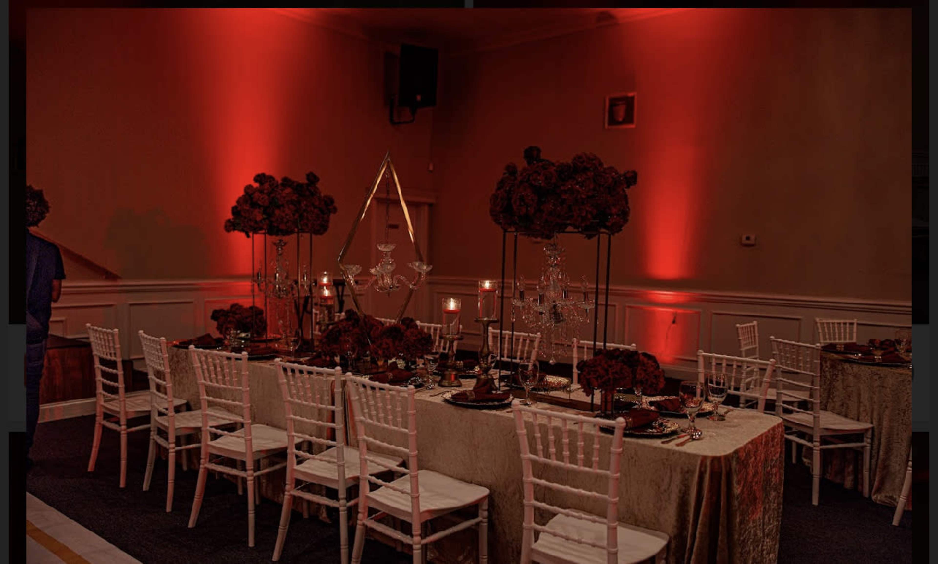 A banquet hall set up for a formal dinner with tables adorned in gold tablecloths, centerpieces of red flowers, and illuminated by soft red lighting.