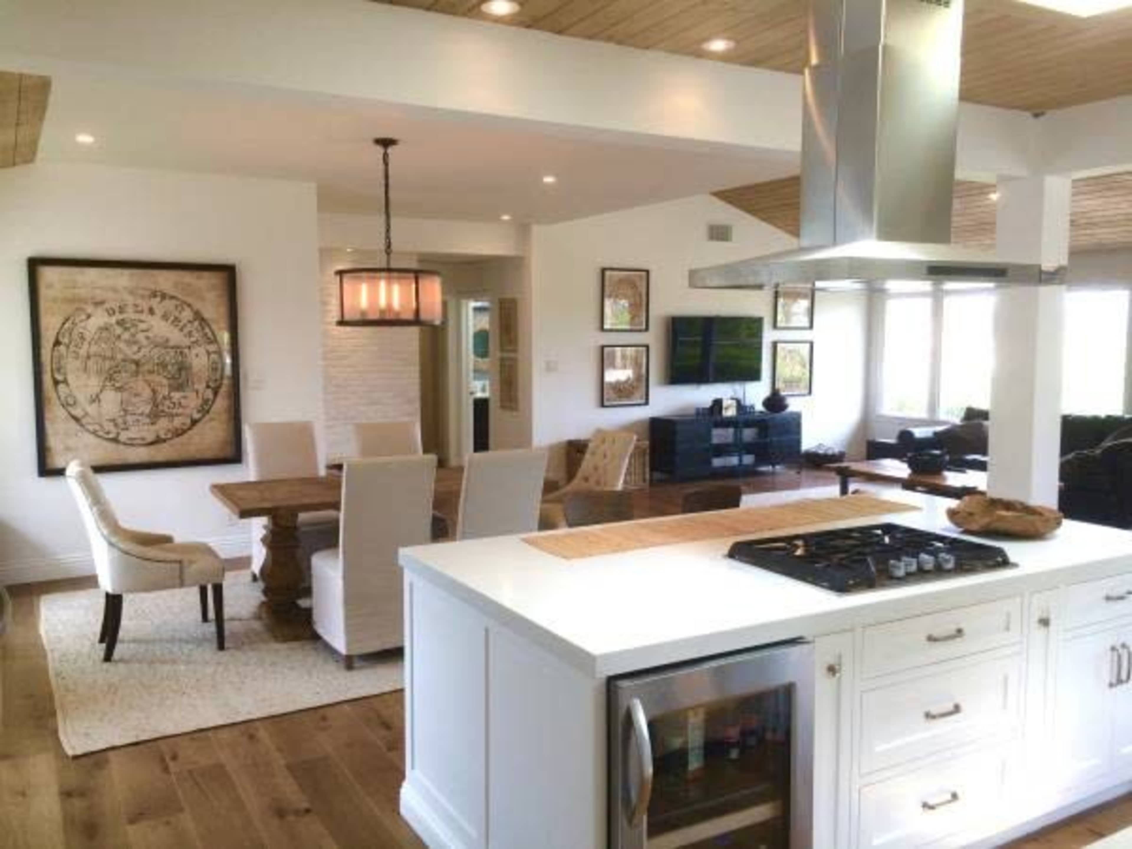 An open-concept kitchen and dining area featuring a white kitchen island, a wooden dining table with chairs, and an adjacent living area with a television and decorative artwork on the walls.