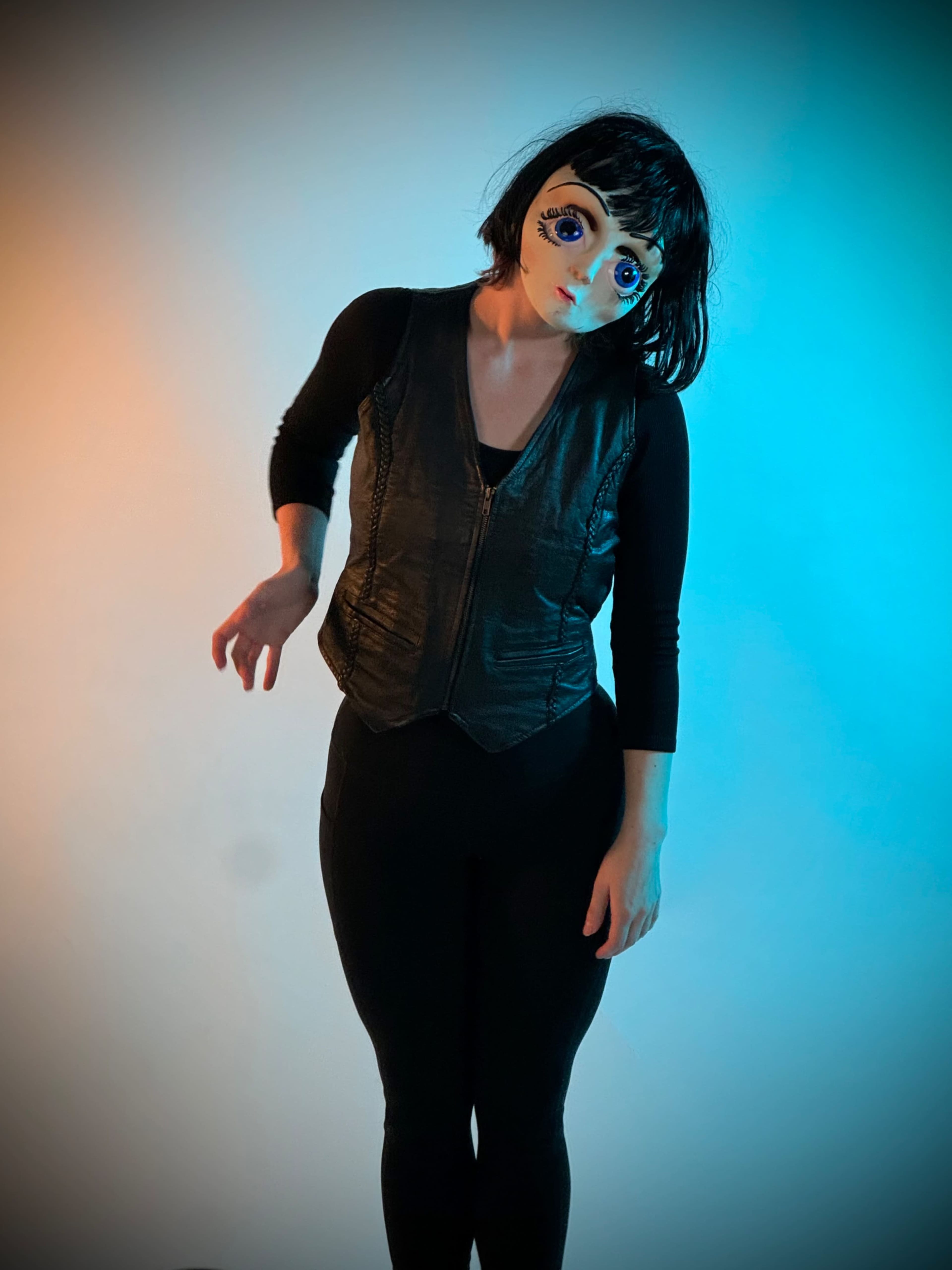 A person wearing a black vest and leggings poses under blue and orange lighting, with a mask featuring large, expressive eyes.