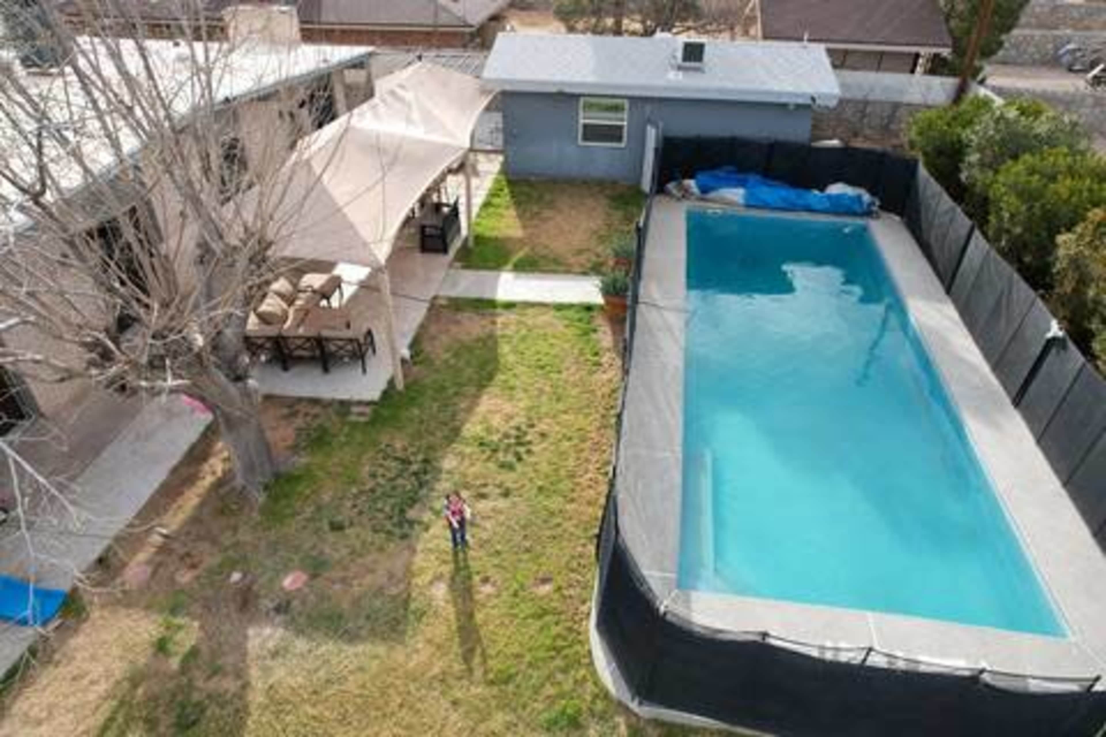 An aerial view shows a backyard with a swimming pool, a shaded seating area, and a house, surrounded by a fence.