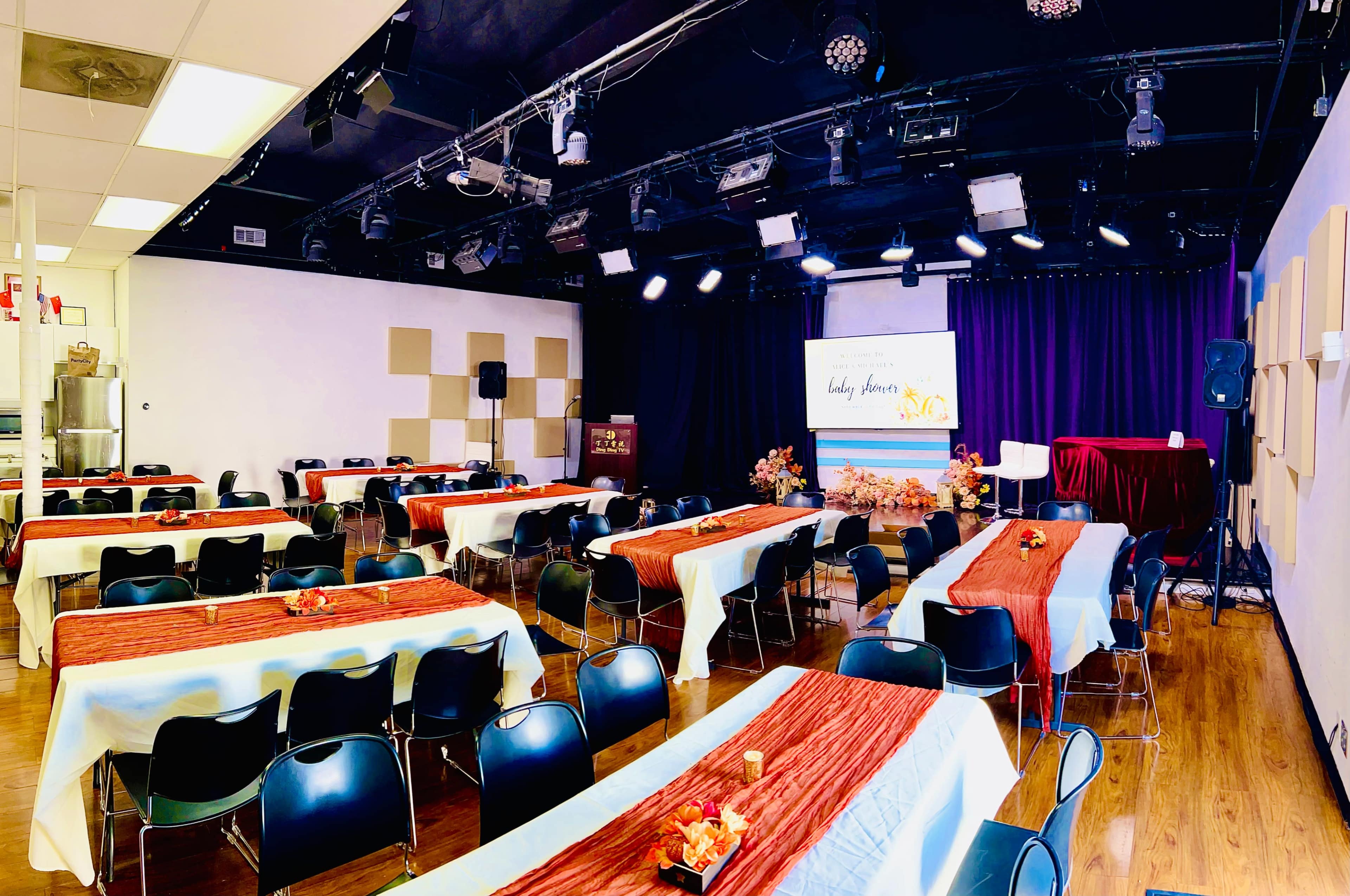 A neatly arranged event space with rows of tables, each covered with white tablecloths and red runners, set for an audience in front of a stage with a projector screen and podium.
