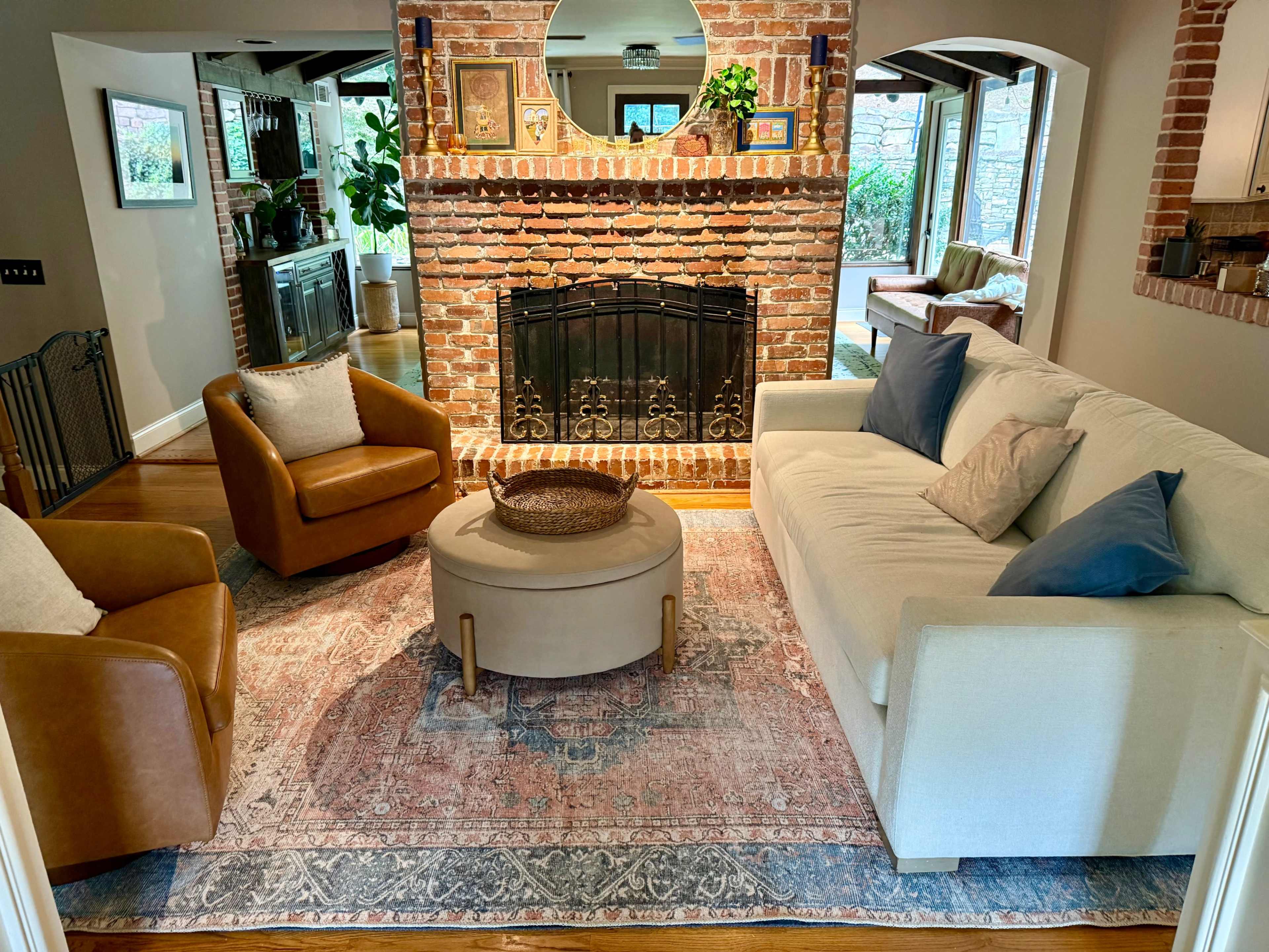 The image shows a living room featuring a brick fireplace, a beige sofa with blue accent pillows, two brown armchairs, and a round coffee table on a patterned rug.