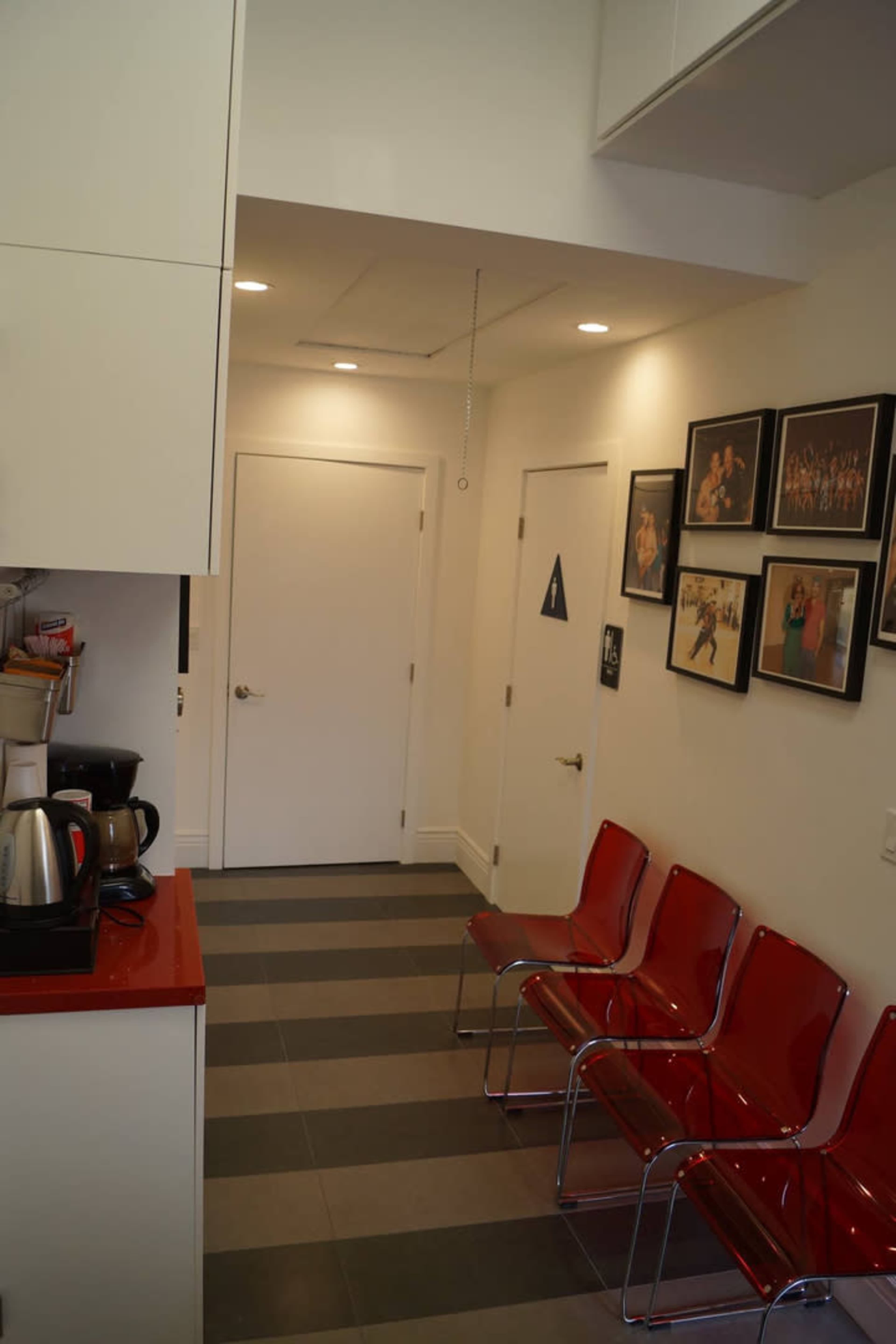The image shows a narrow interior space featuring a kitchenette area on the left, a closed door at the end of the hallway, and rows of red chairs along the right wall, adorned with framed photographs.
