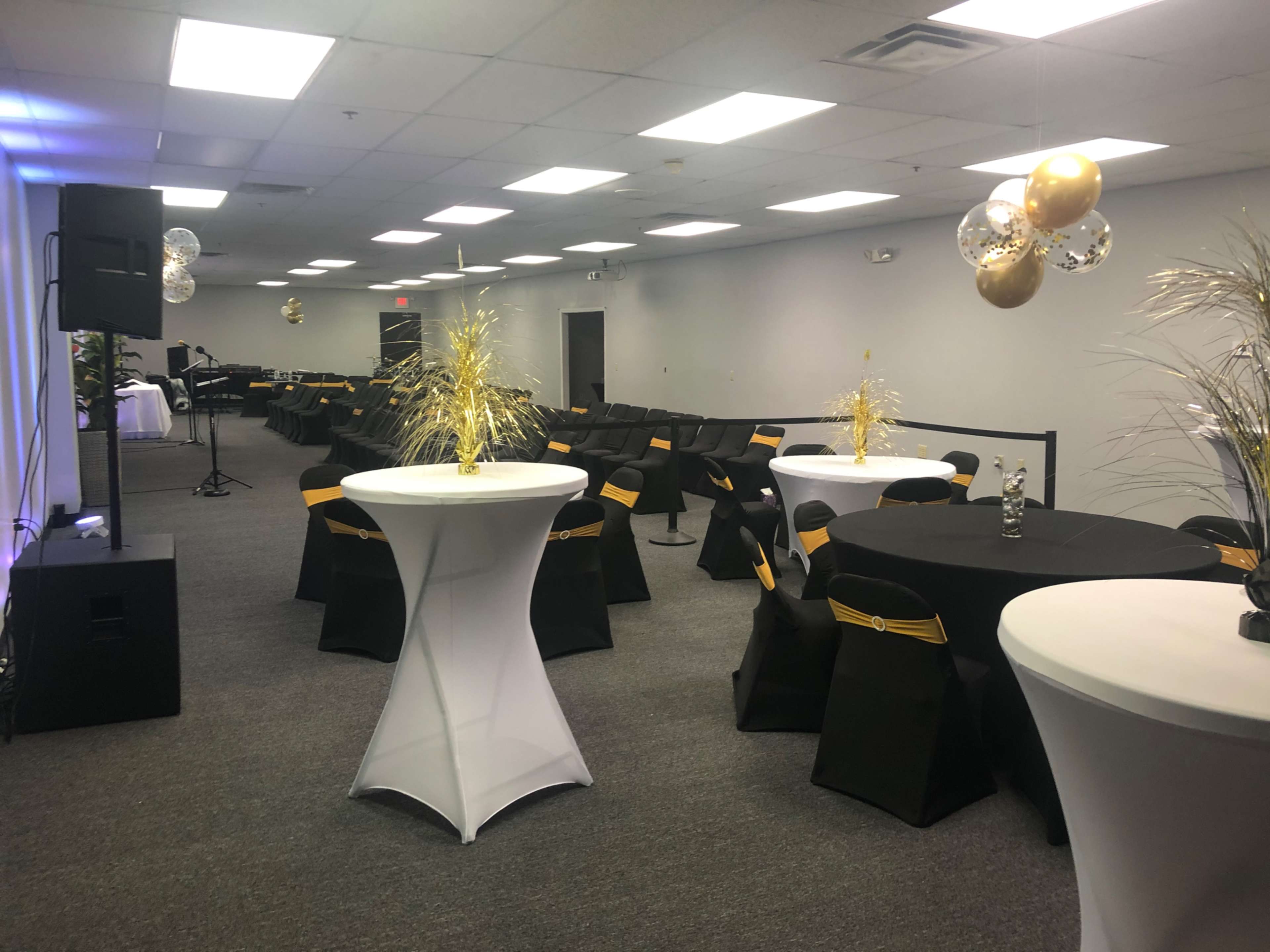 The image shows a banquet hall set up for an event, featuring round black and white tables with gold accents and decorations, alongside a stage with sound equipment.