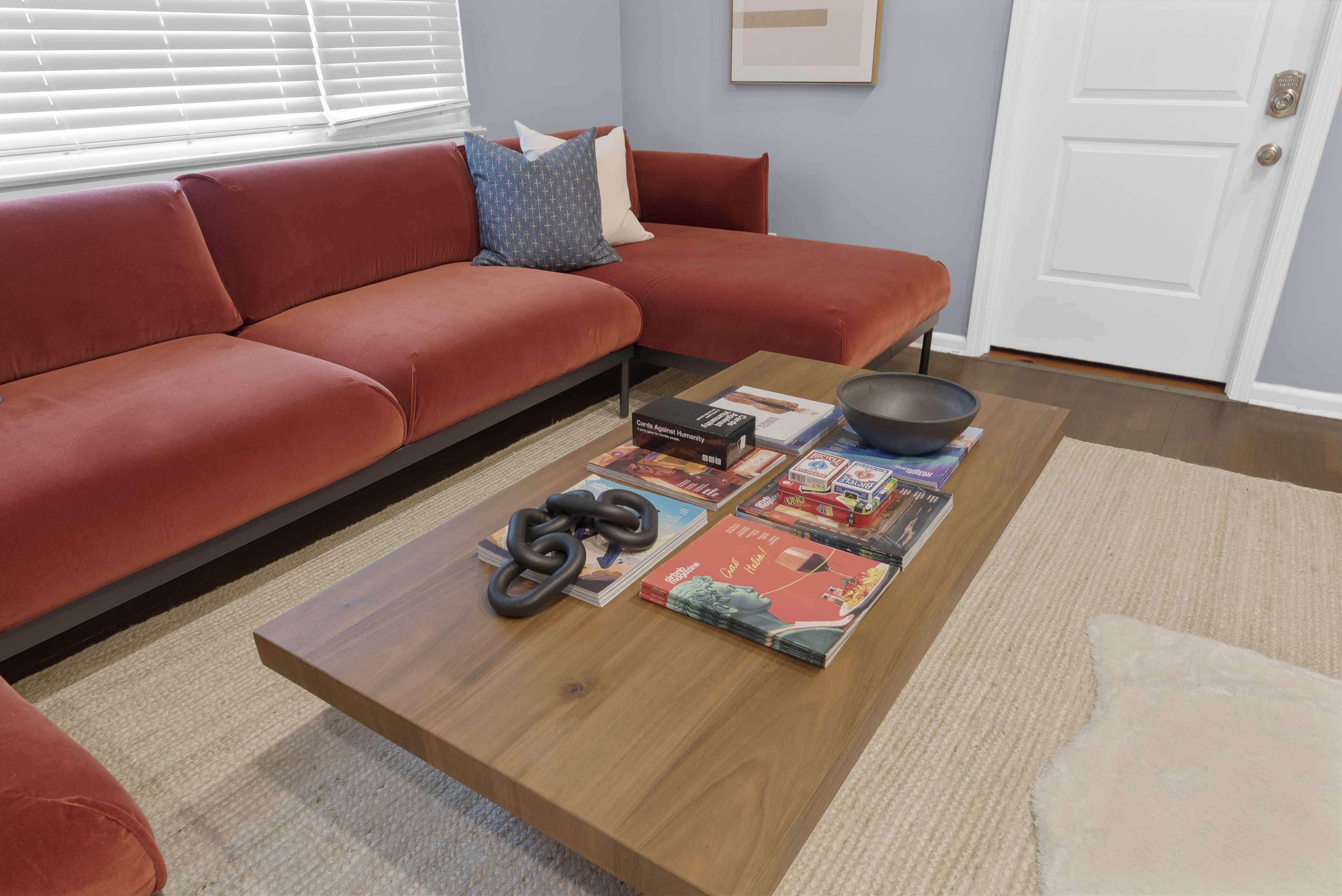A modern living room features a red sectional sofa, a wooden coffee table stacked with books and decorative items, and a light-colored area rug.