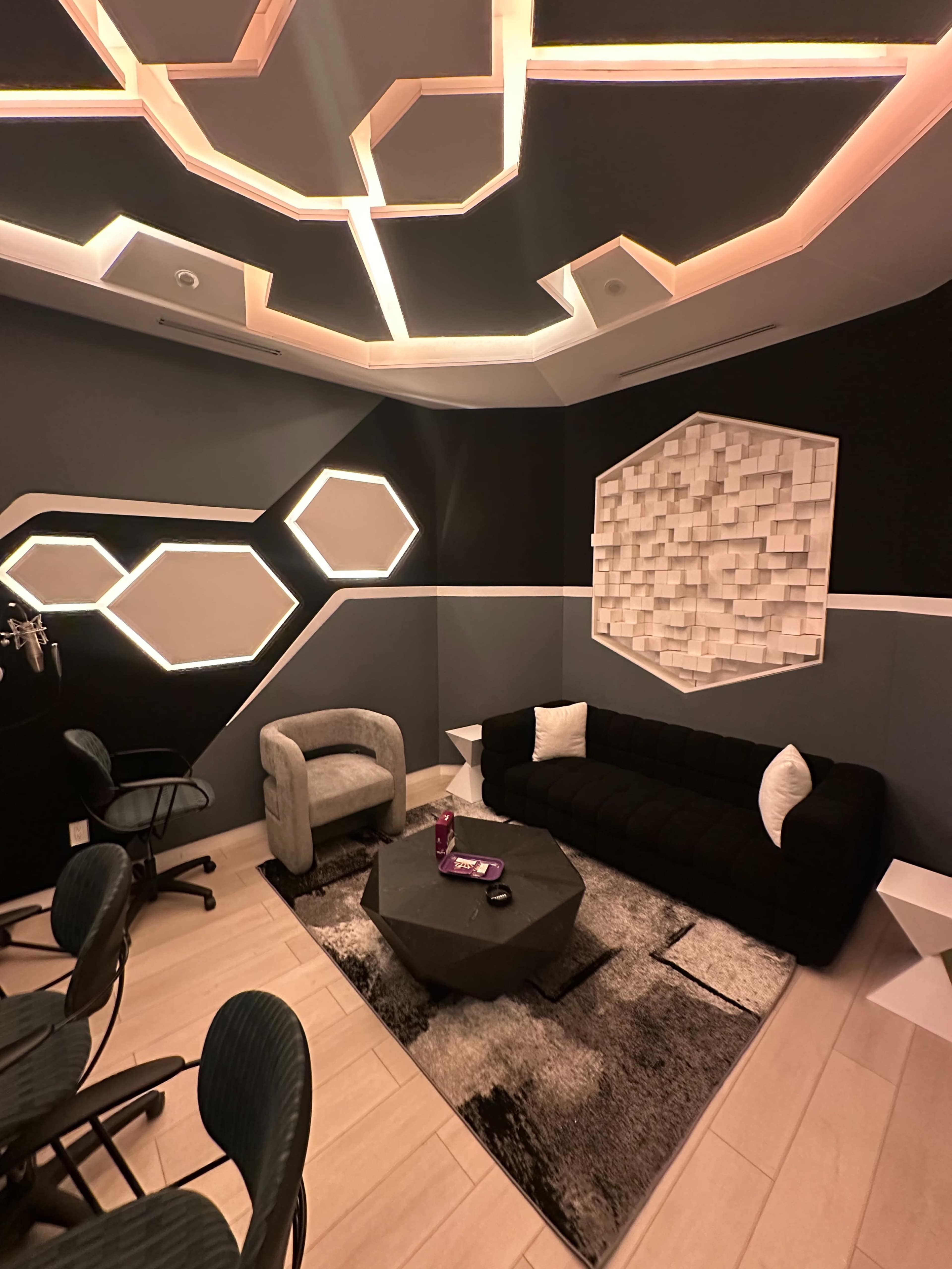 The image shows a modern lounge area with a black sofa, a light gray armchair, a geometric wall design, and soft lighting.