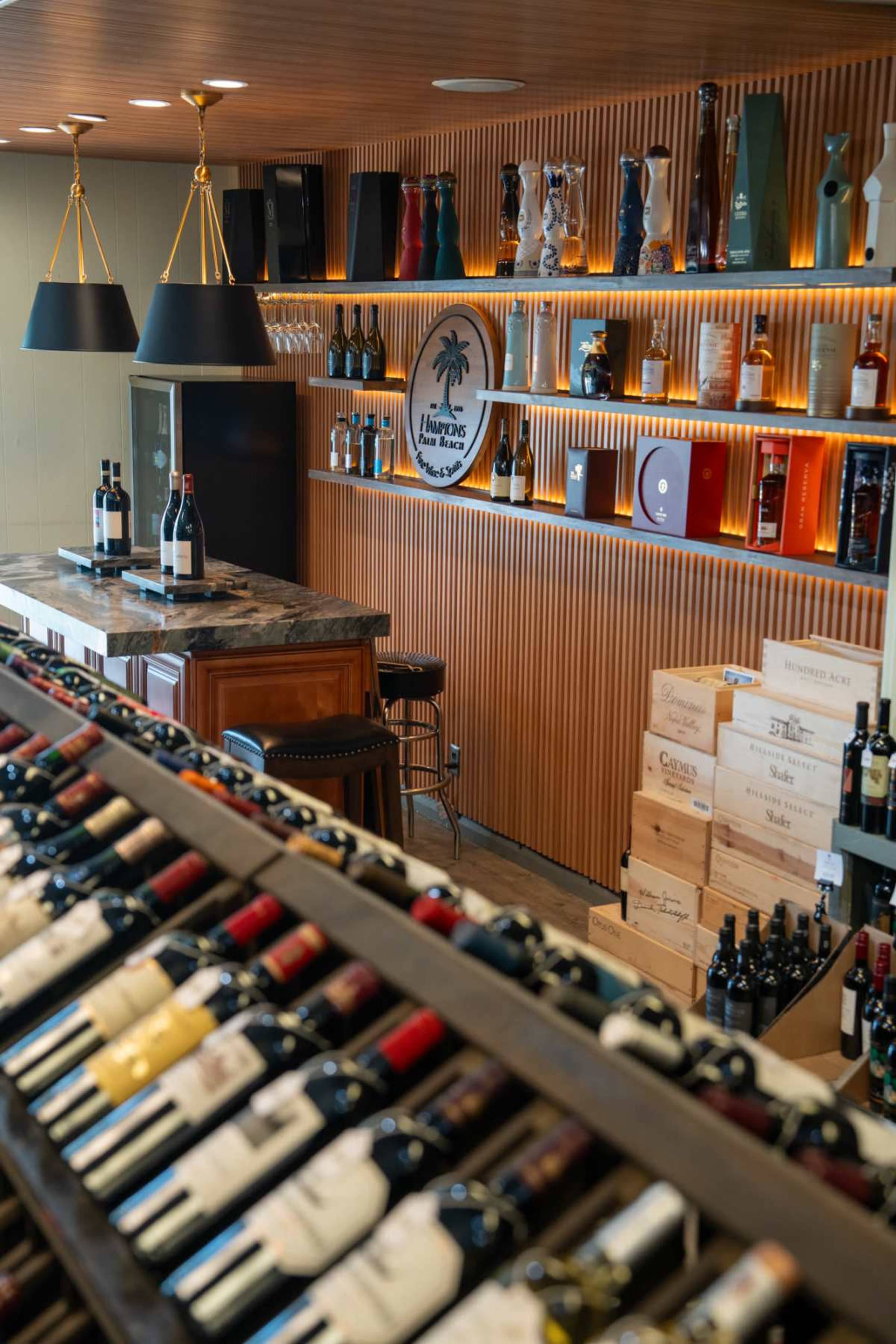 The image shows a wine bar with a display of wine bottles in the foreground and a polished countertop with various spirits and decor in the background.