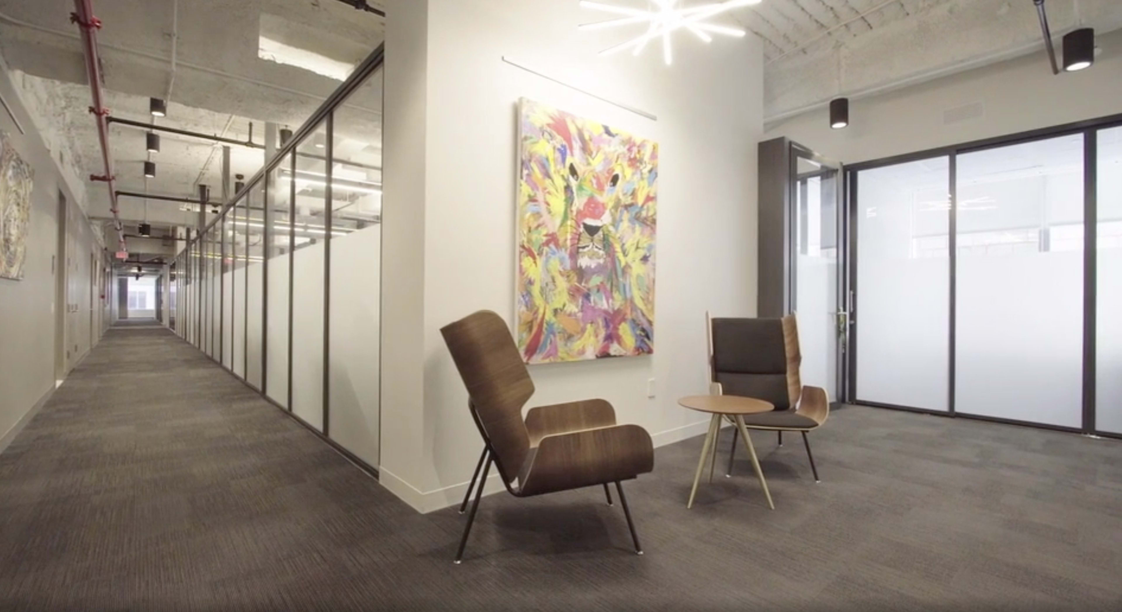 A modern office space with a corridor lined with glass-walled cubicles and a seating area featuring two chairs and a small table.