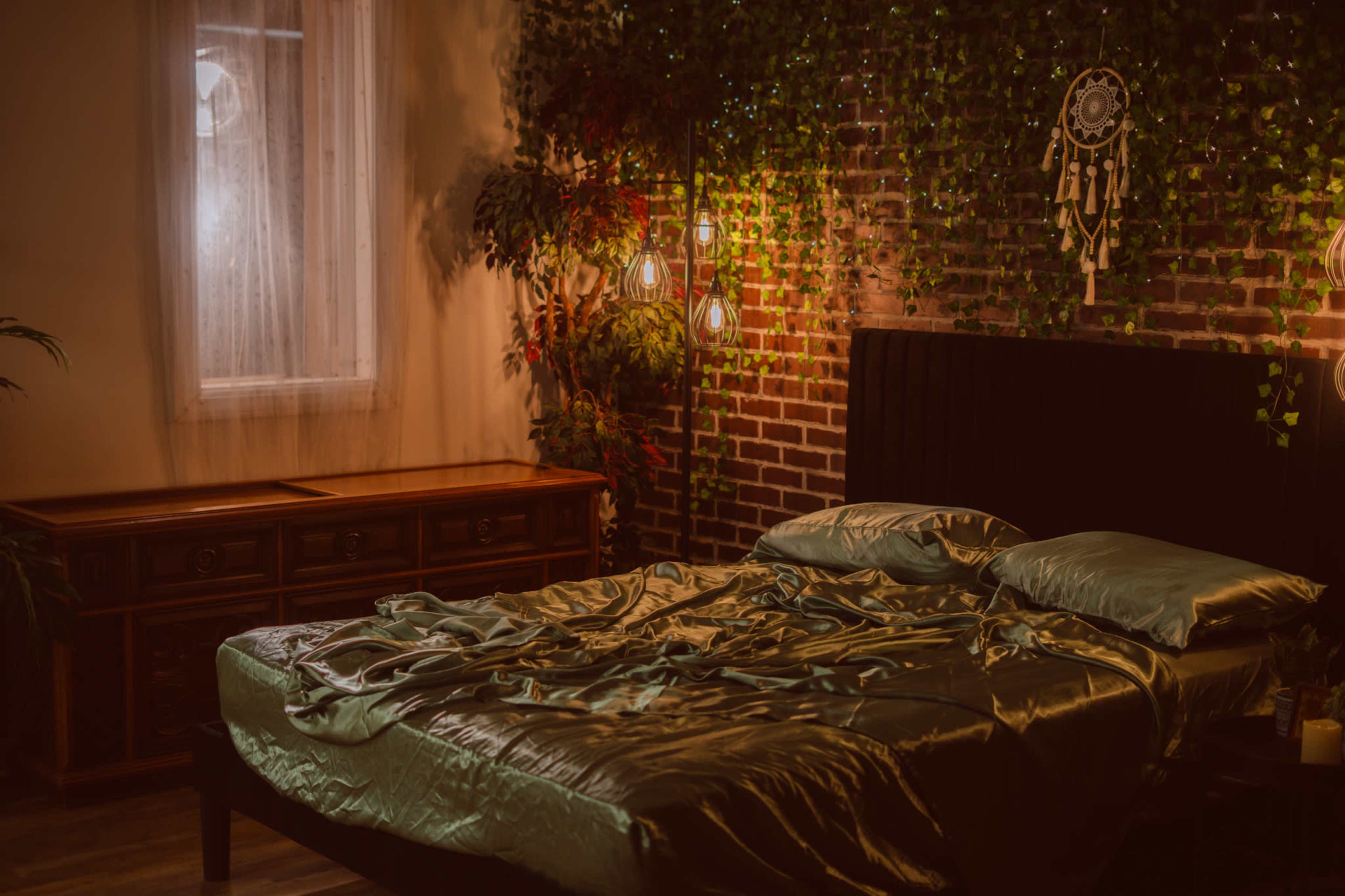 The image shows a bedroom with a large, unmade bed covered in green satin sheets, adjacent to a wooden dresser, and decorated with hanging plants and soft lighting.