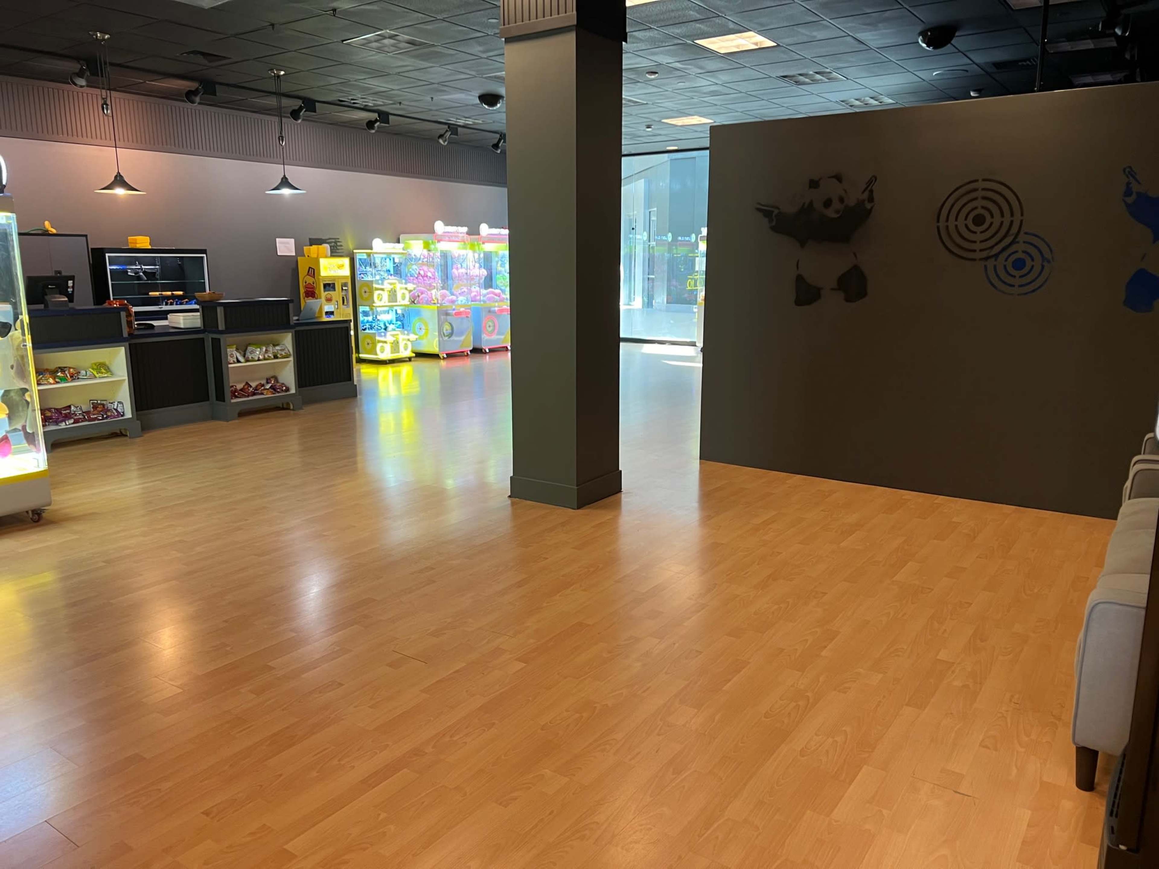 The image shows an empty indoor space with wooden flooring, featuring arcade machines in the background and a partition wall on one side.