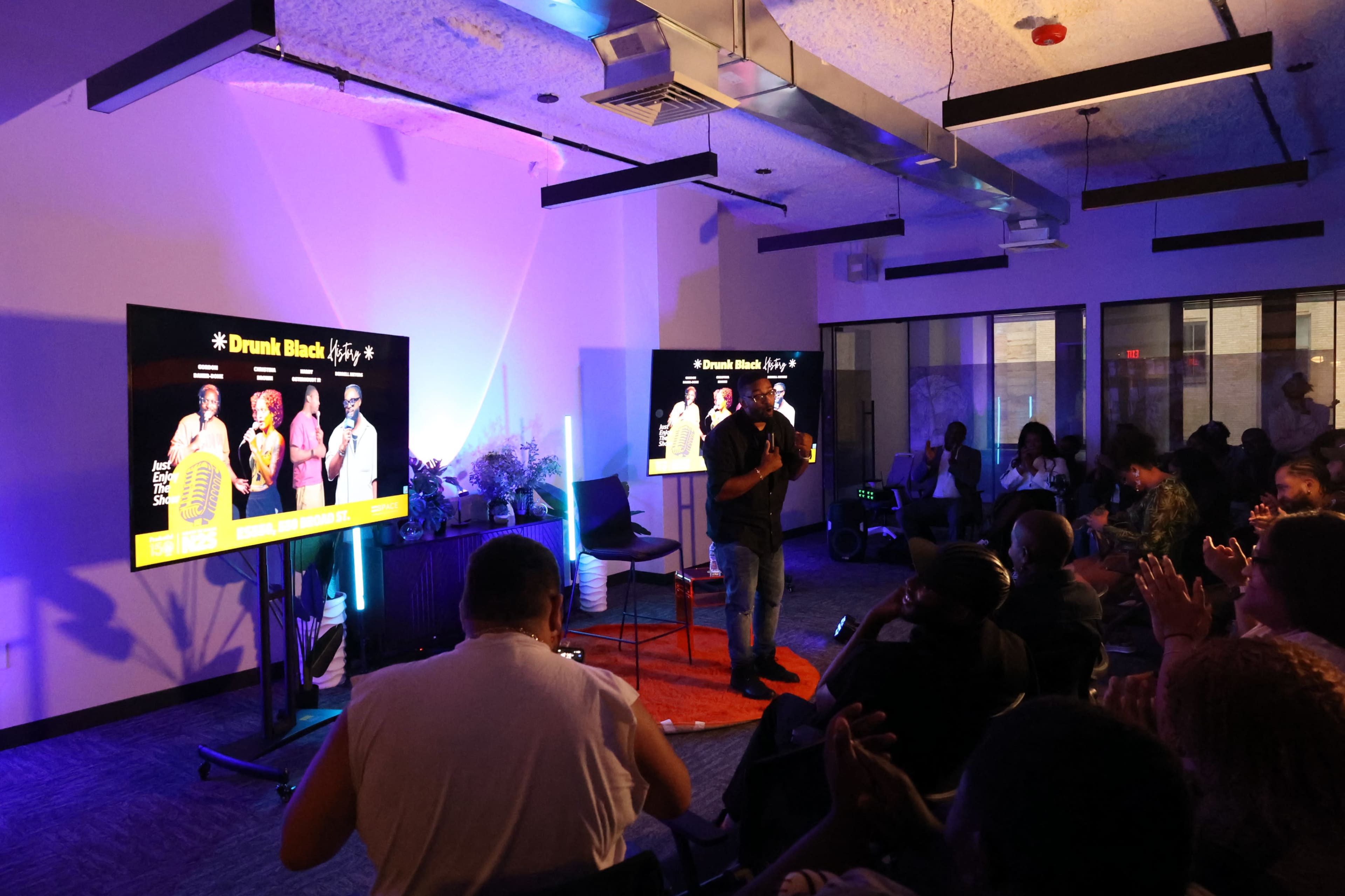 A speaker stands on stage in front of an audience, with large screens displaying colorful images and text.