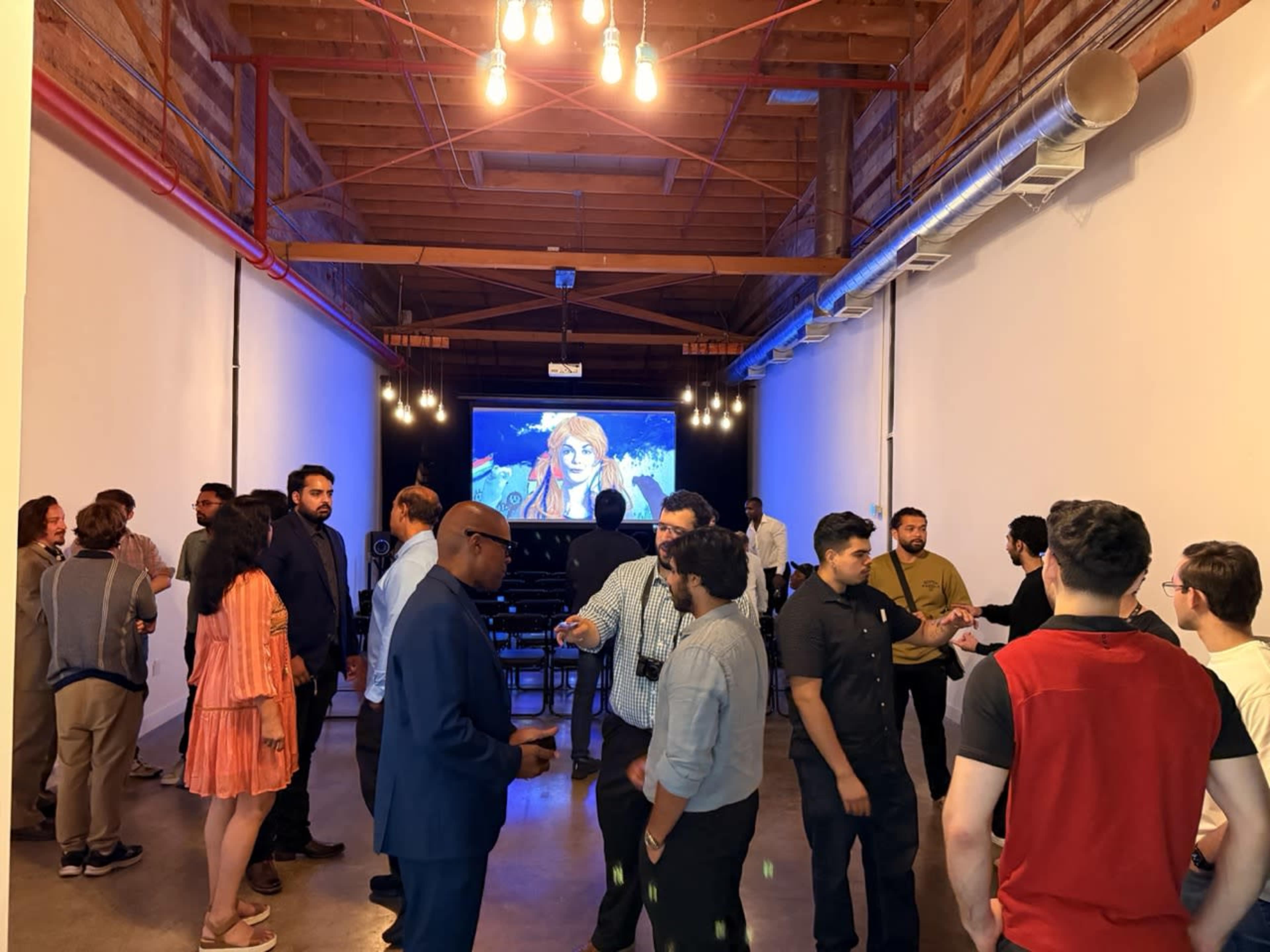 A group of people is socializing in a modern event space with a large screen displaying an image in the background.