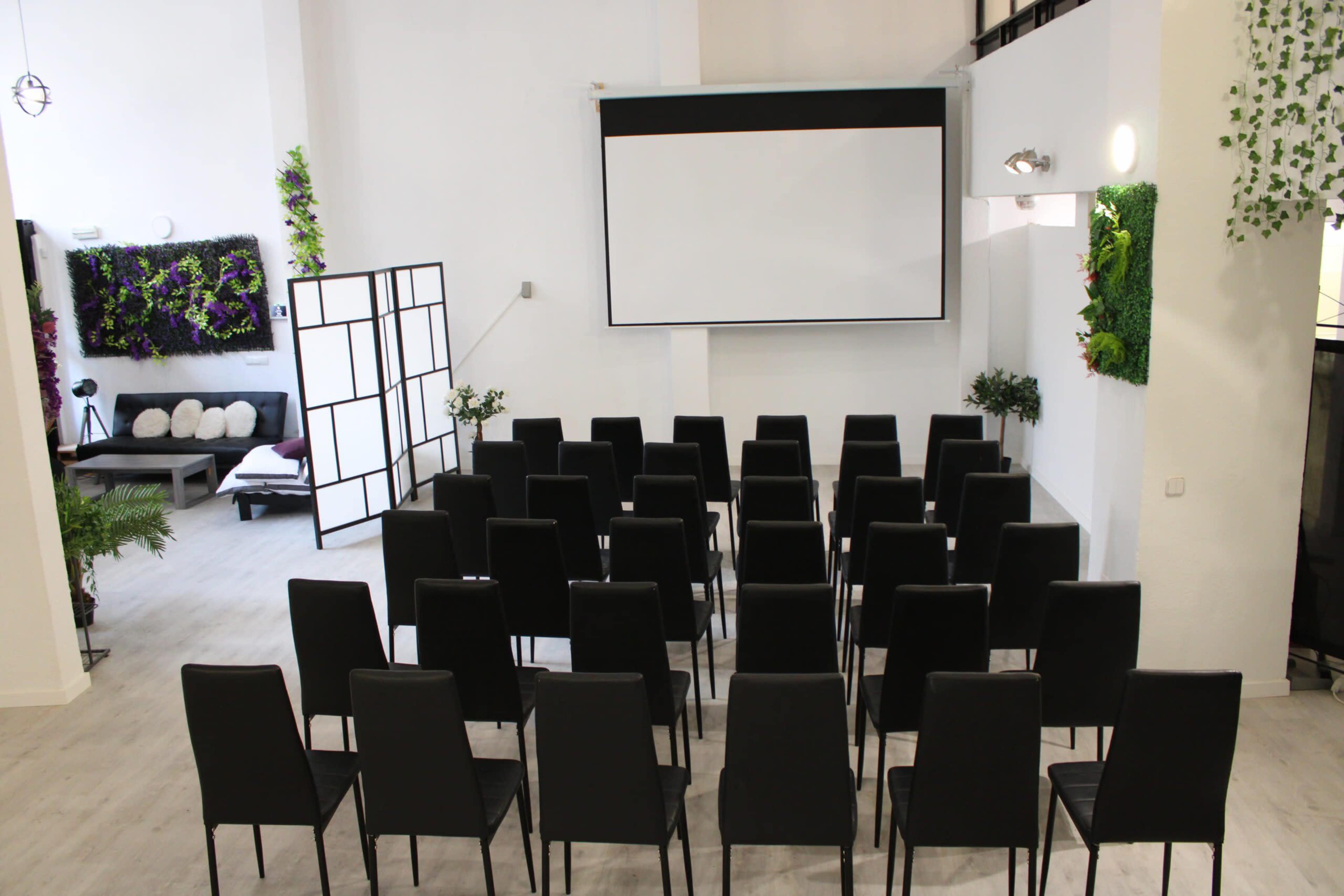 The image shows a minimalist room set up with rows of black chairs facing a large projection screen, with greenery decor on the walls.