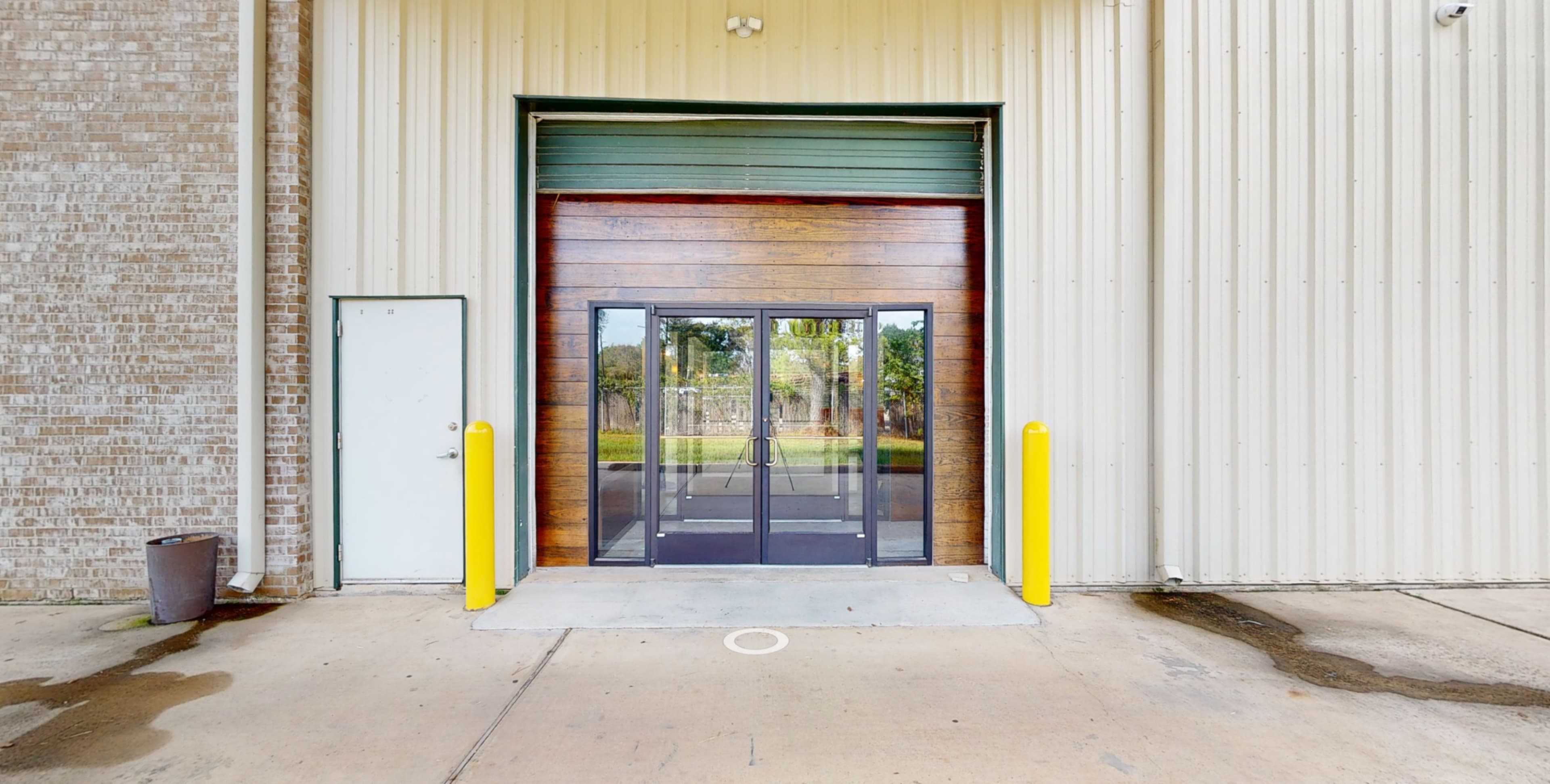 A large glass door set in a wooden frame, flanked by yellow bollards, leading into a metal-clad building.