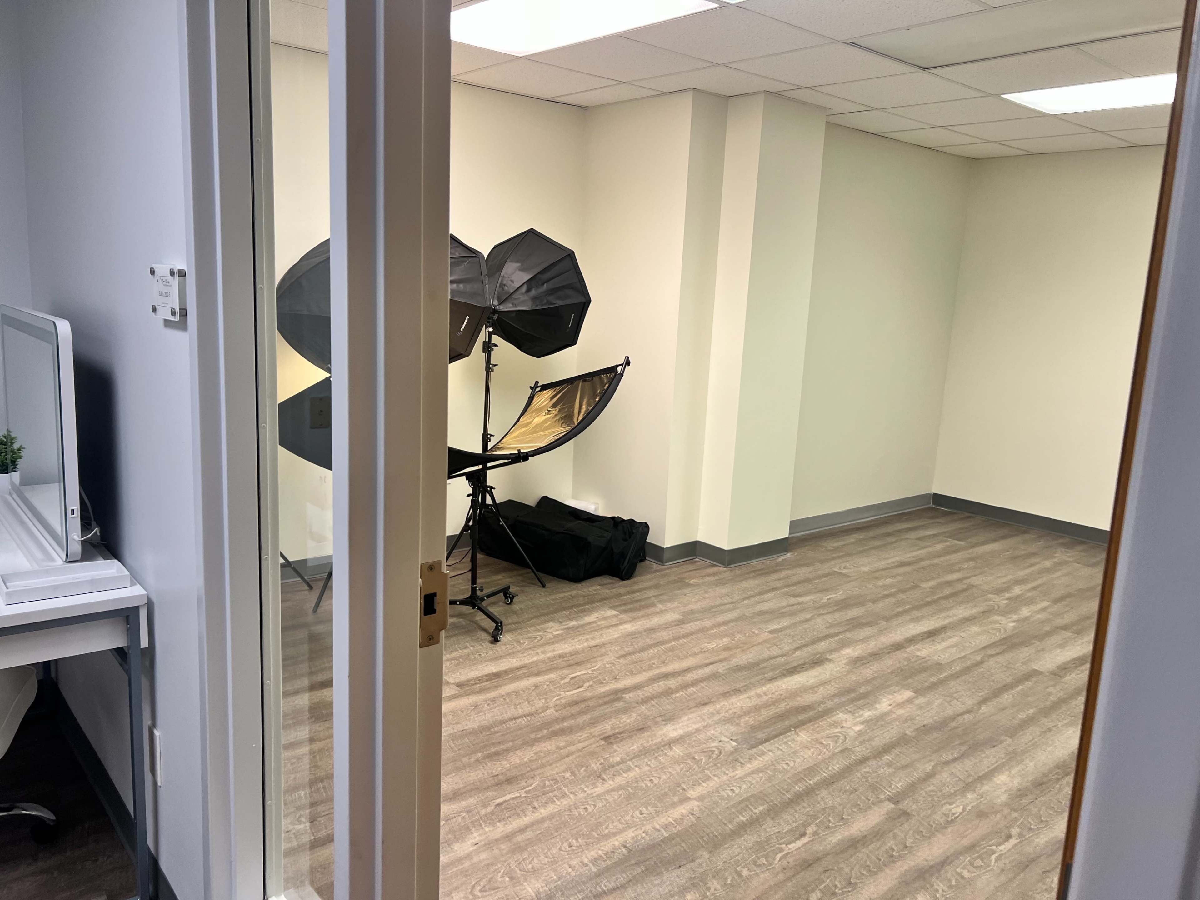 A well-lit empty room features photography equipment, including setups for lighting, alongside a small desk with a mirror.