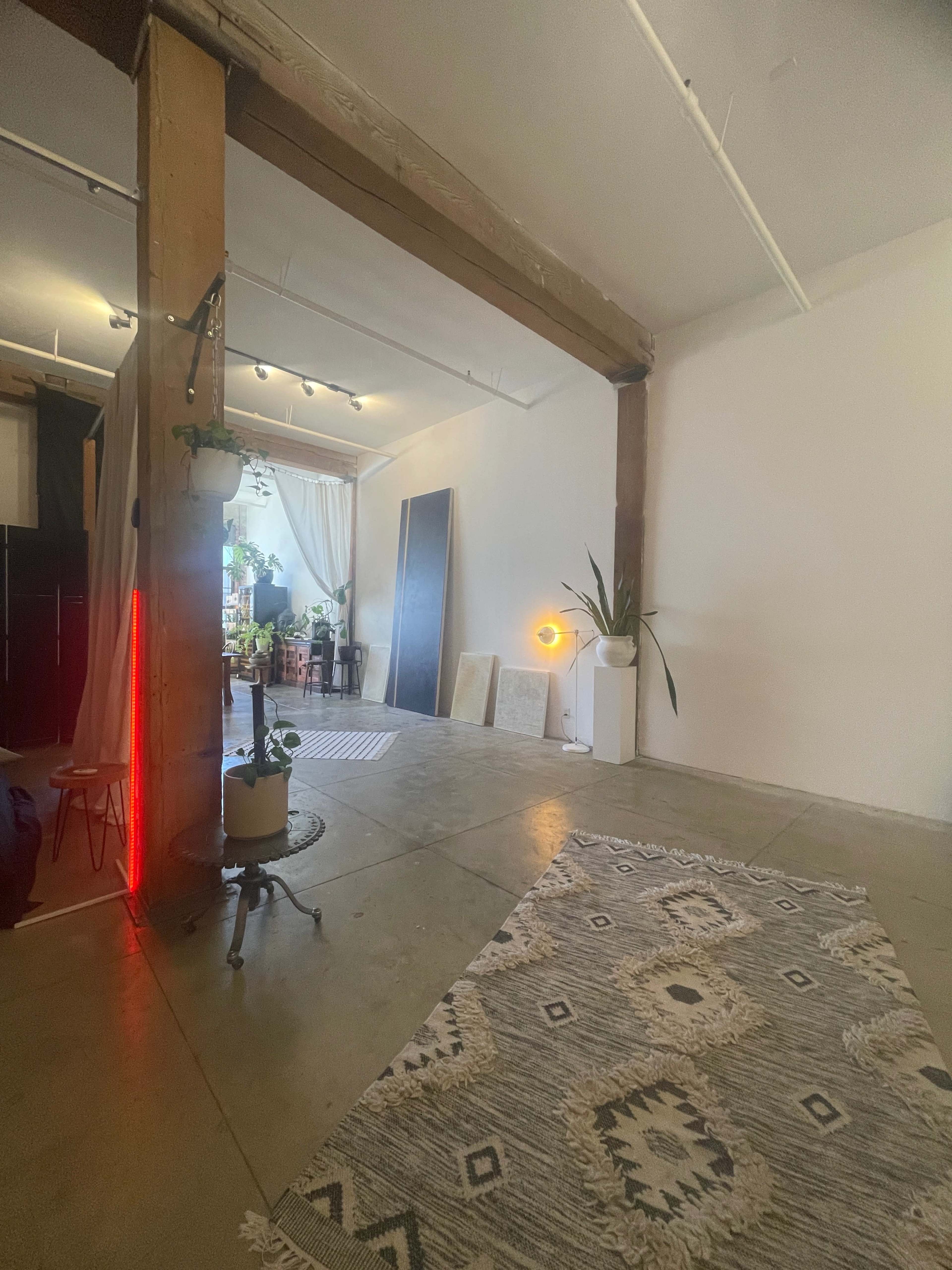 A spacious room with concrete flooring, featuring a patterned rug, a plant, and a warm light source, along with large windows draped with sheer curtains.