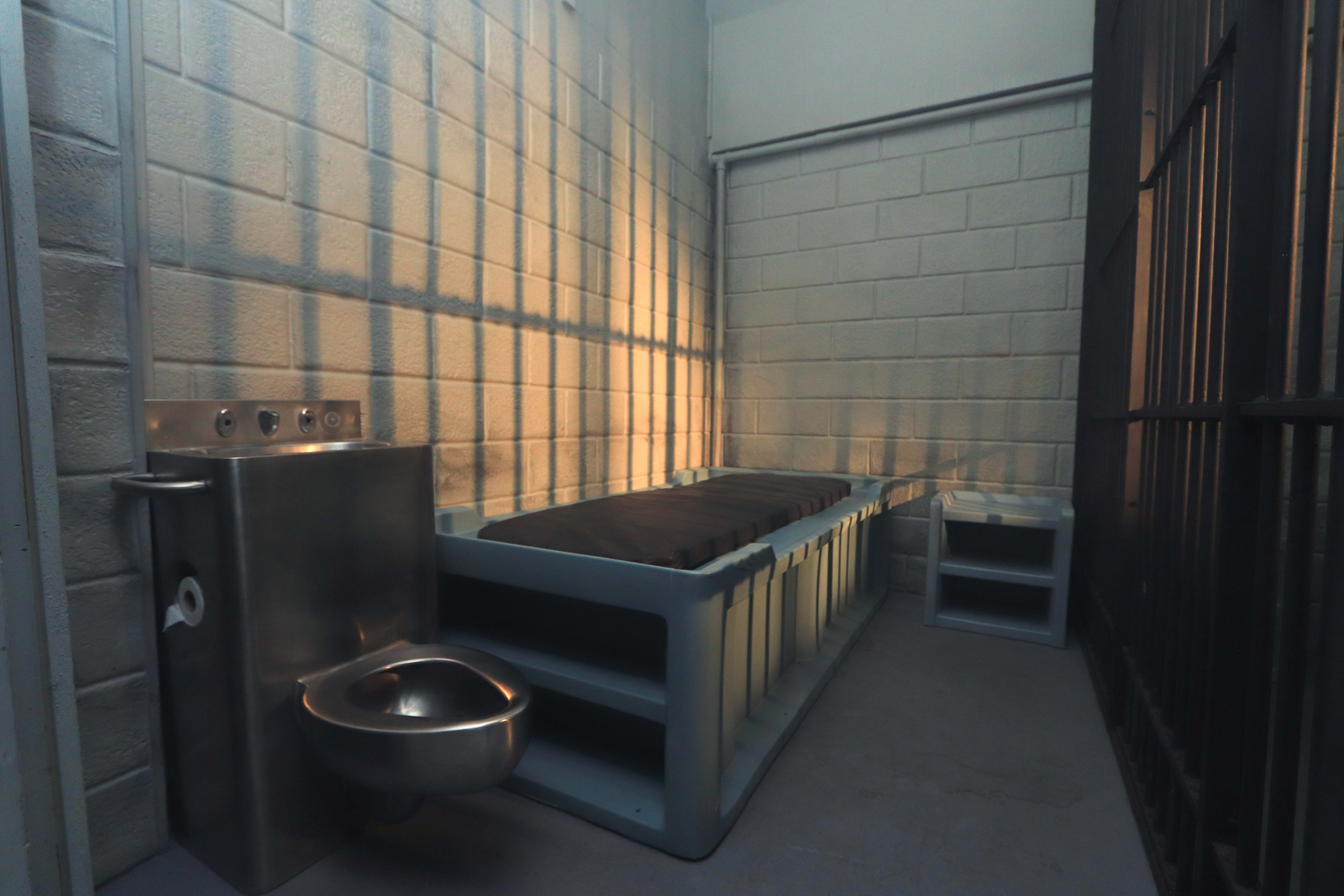 The image shows a small, stark prison cell with a bed, a metal toilet, and a small shelf, illuminated by slanted light from a barred window.