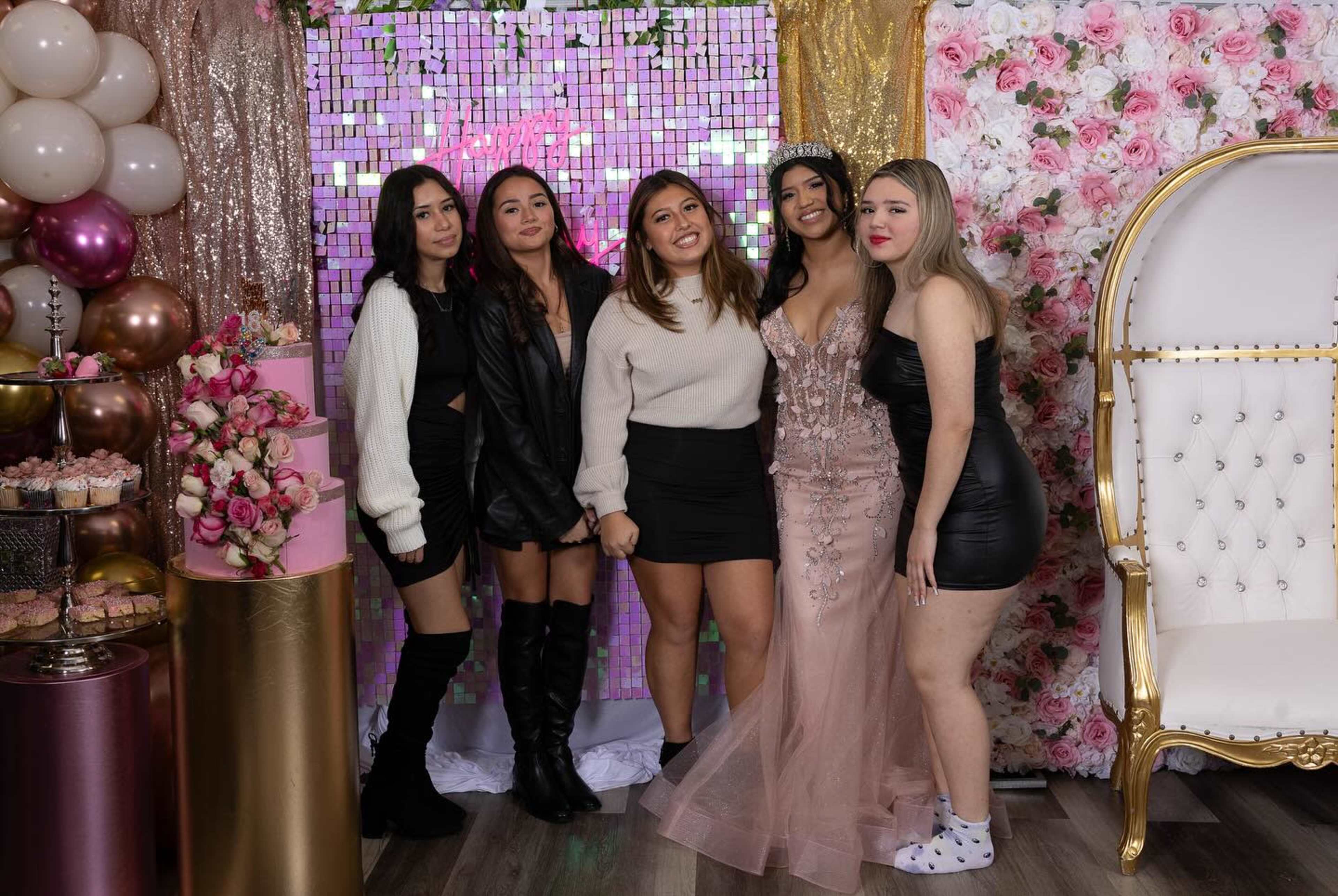 A group of five women poses together in a decorated party area featuring balloons, a floral backdrop, and a cake display.