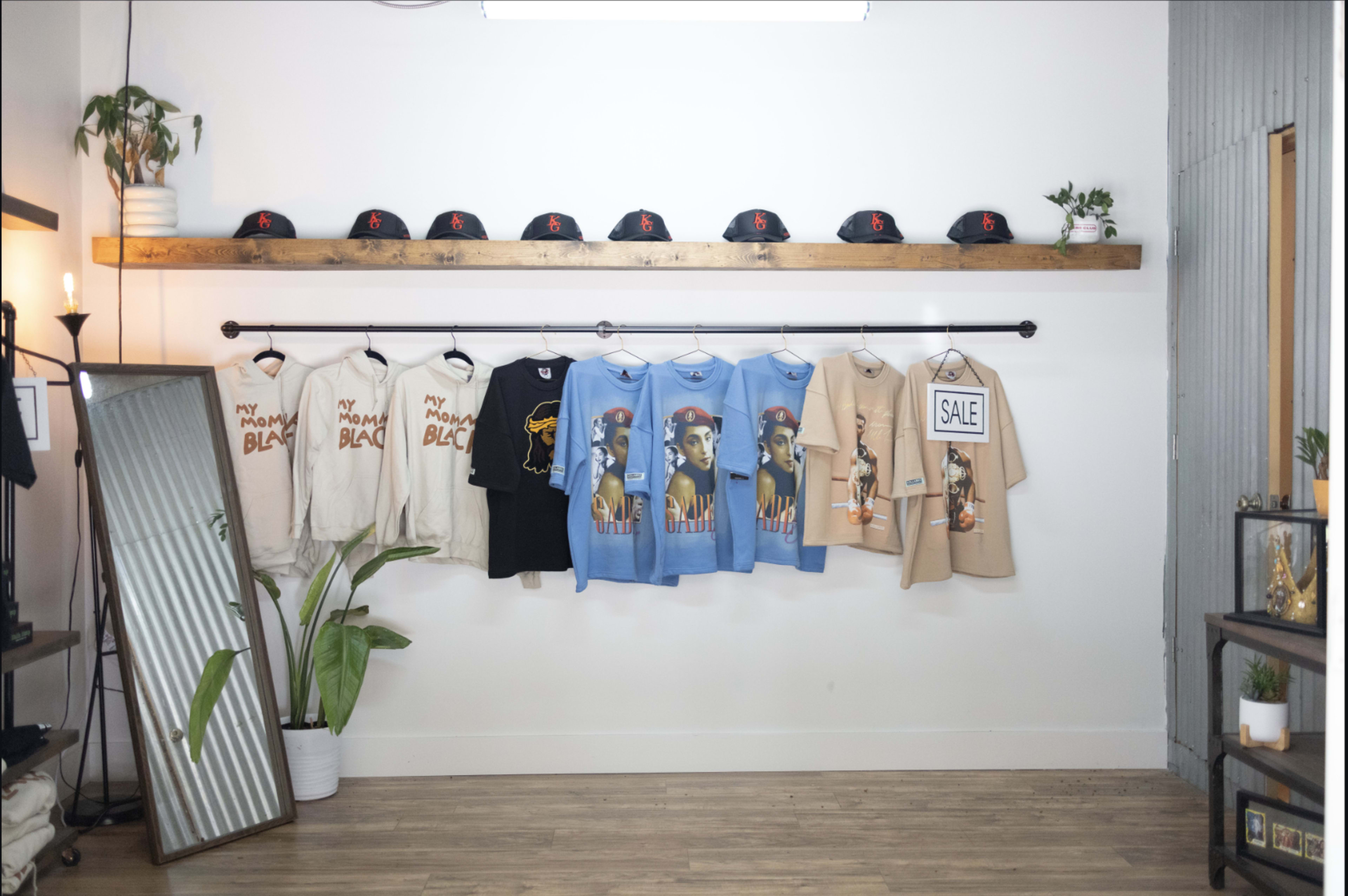 The image shows a clothing display featuring t-shirts and hats arranged on a wall with a mirror and decorative elements in a boutique setting.