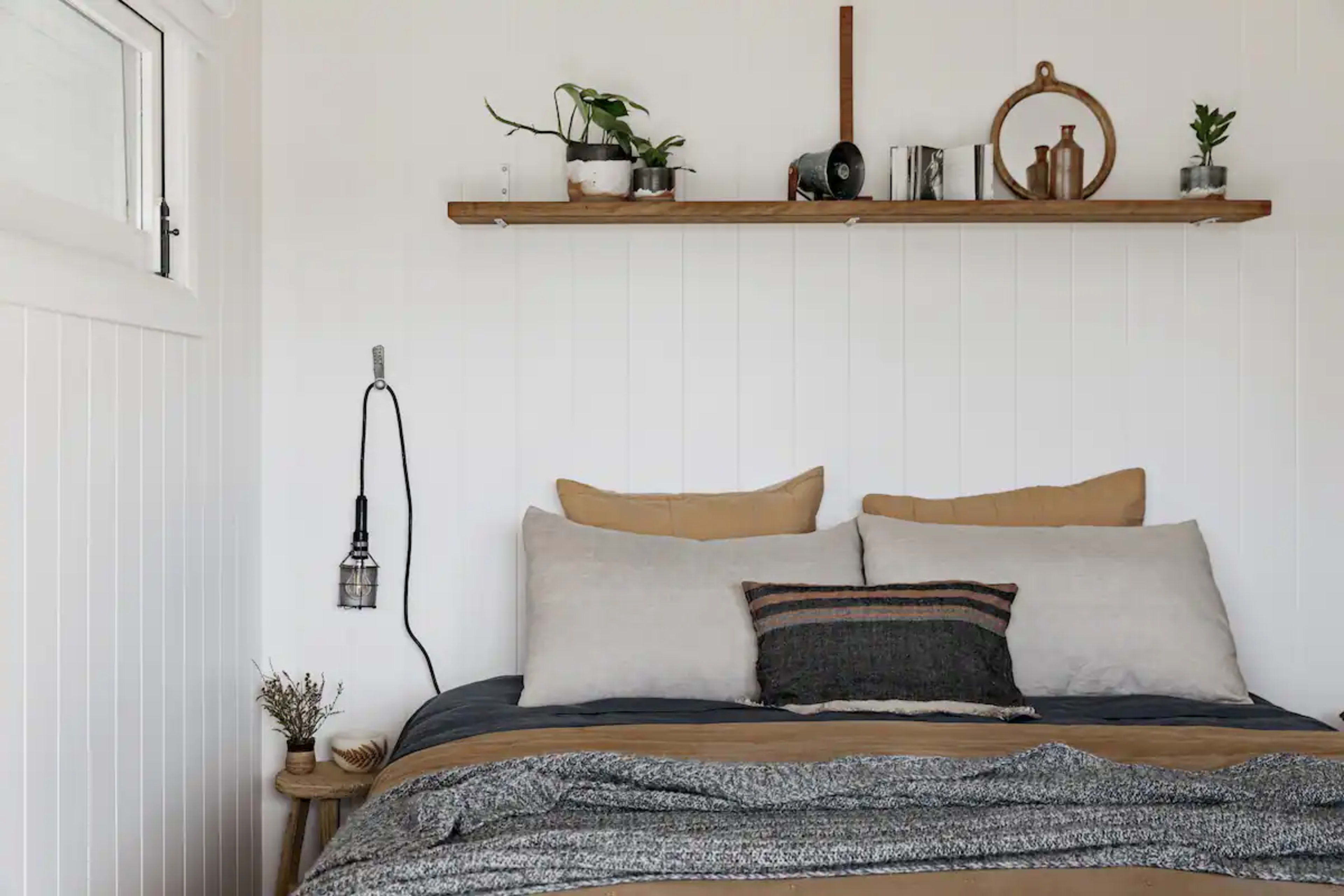 A neatly made bed with multiple cushions, a wooden shelf above adorned with potted plants and decorative items, and a wall-mounted light fixture.