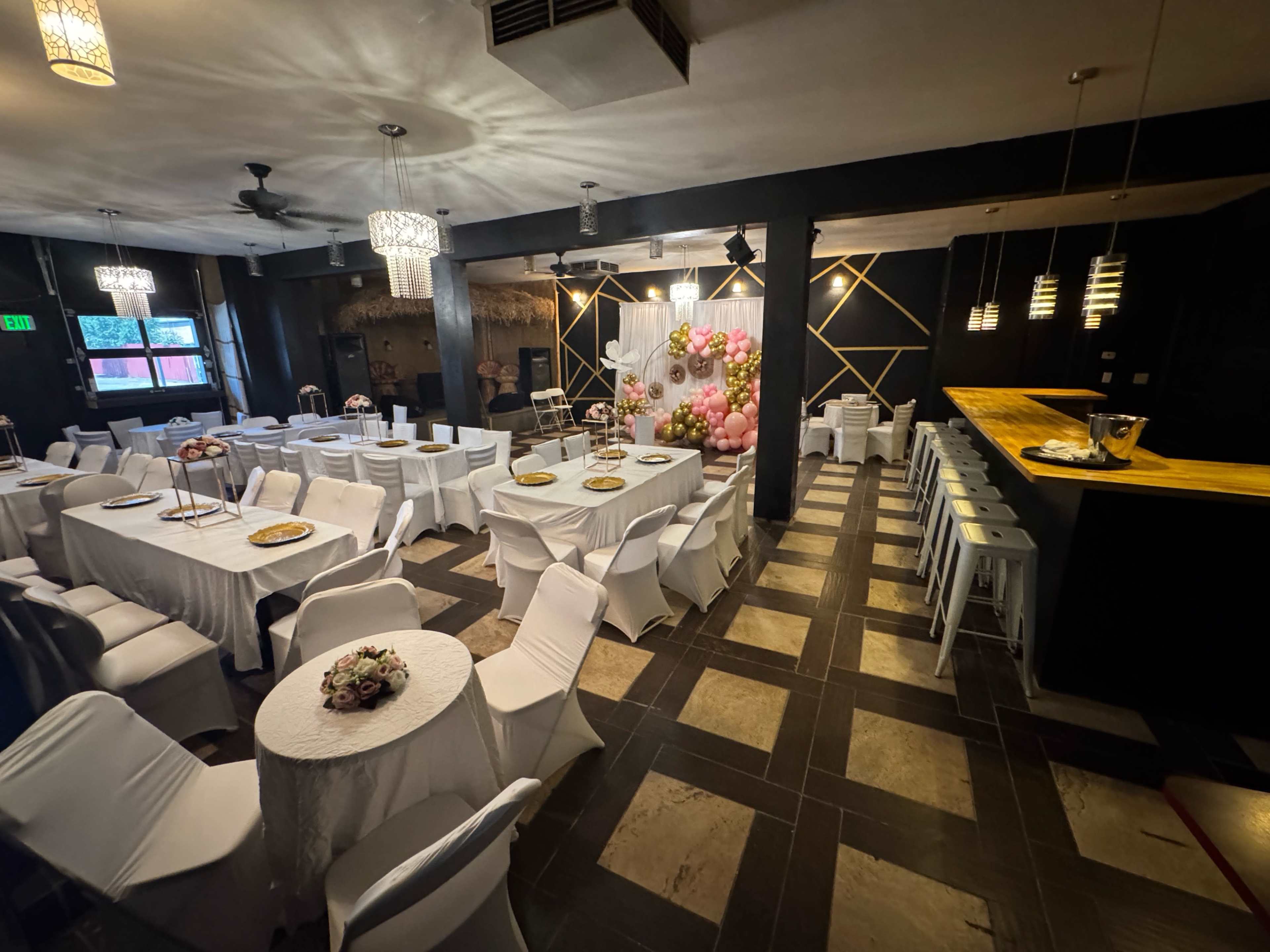 The image shows a modern event space with white tablecloths, chairs, and a decorative backdrop featuring pink balloons.
