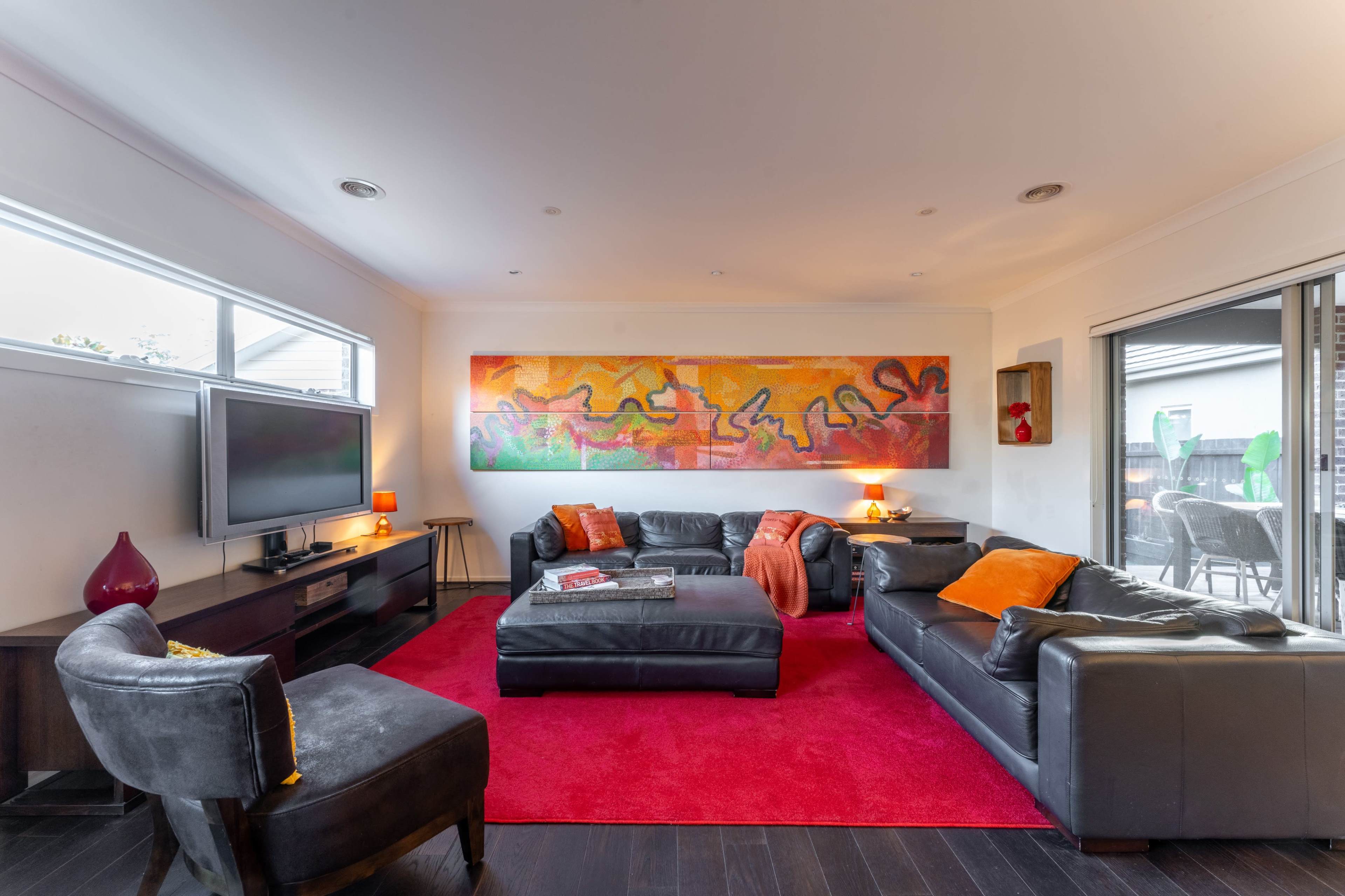 A living room features dark leather furniture, a large colorful artwork on the wall, and a red area rug.