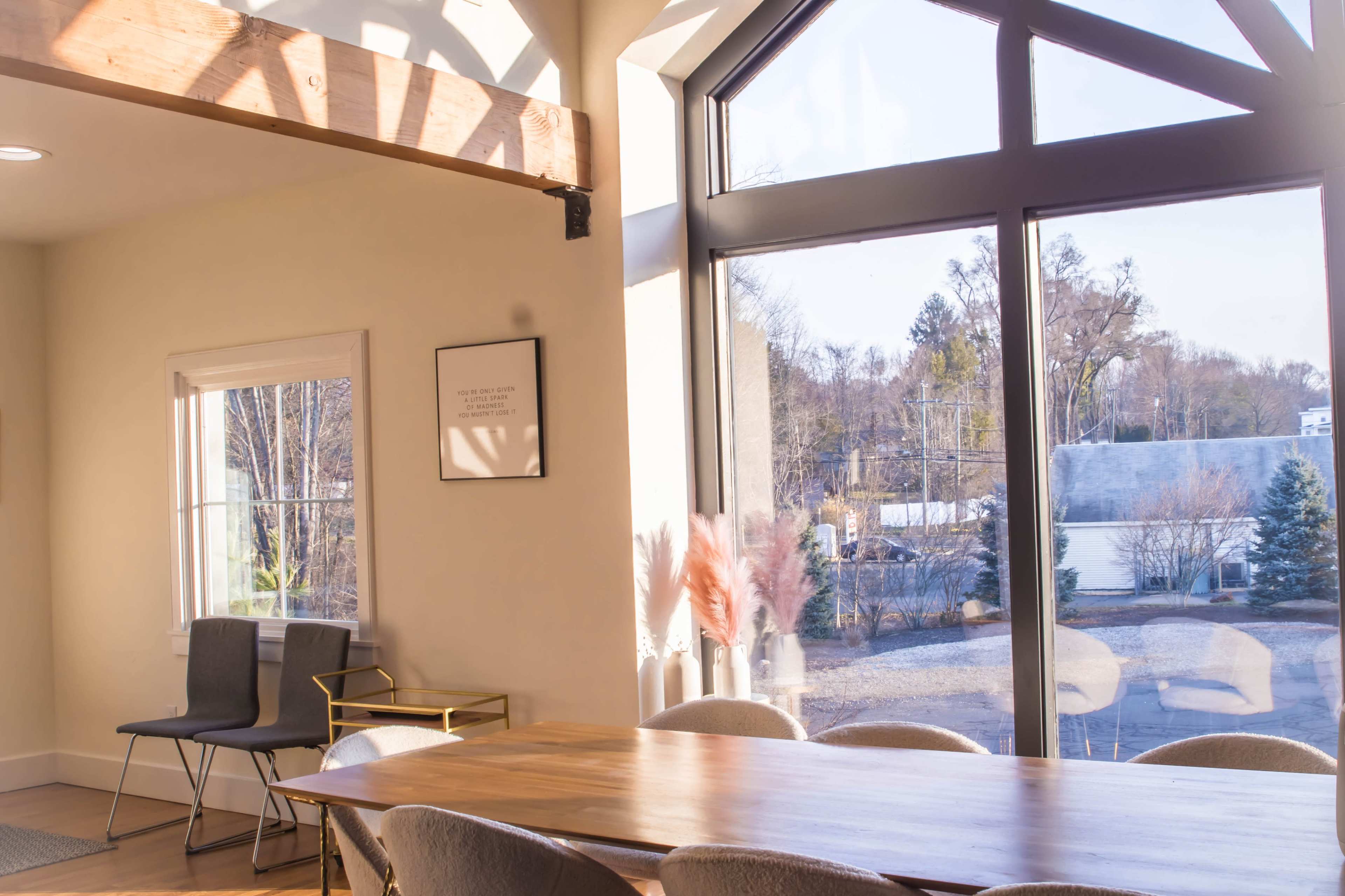 A spacious room features a large window with natural light, a wooden table, and chairs arranged around it.
