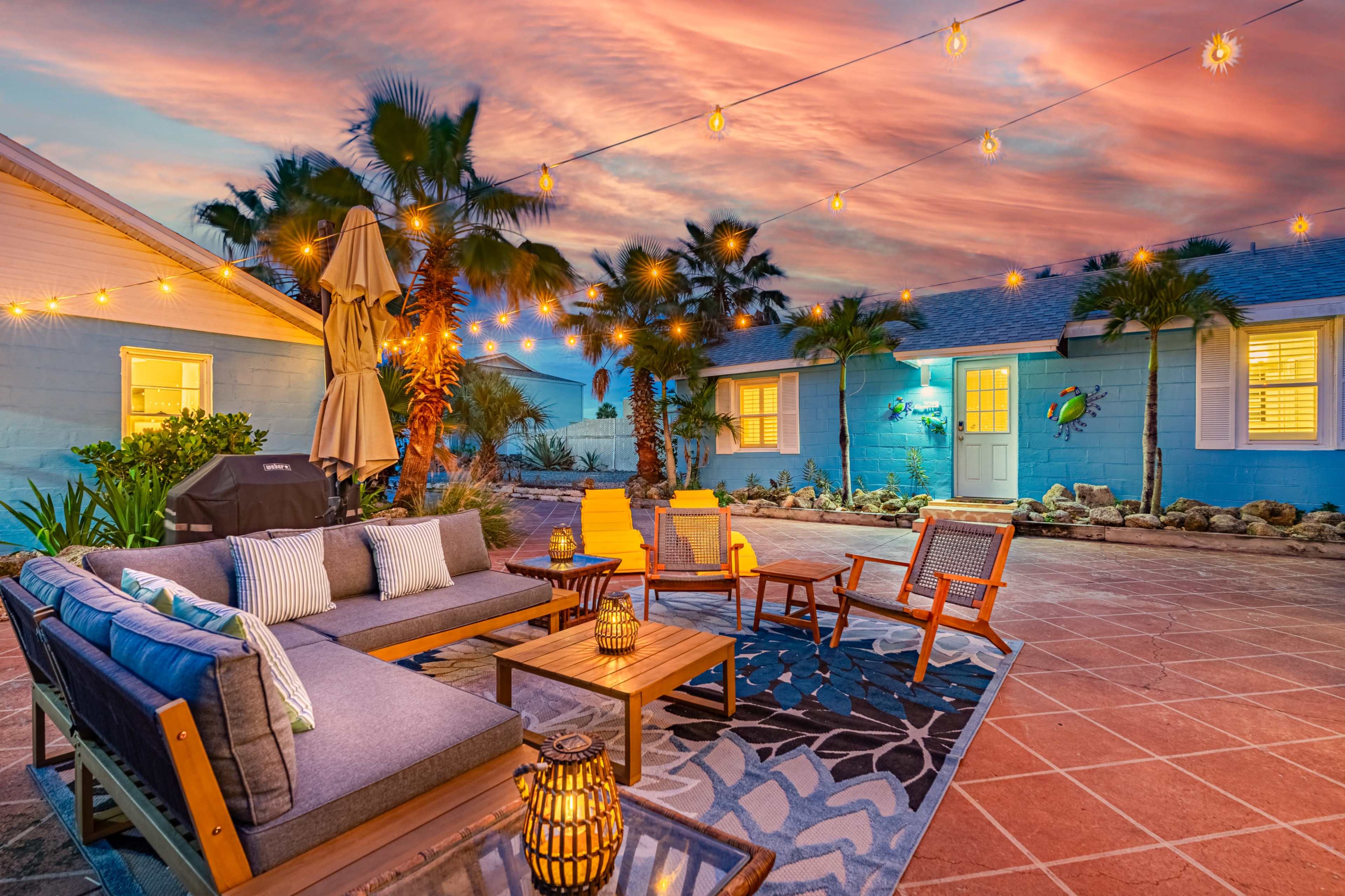 A cozy outdoor seating area features a gray sofa, wooden chairs, and string lights, set against a backdrop of palm trees and a colorful sunset sky.