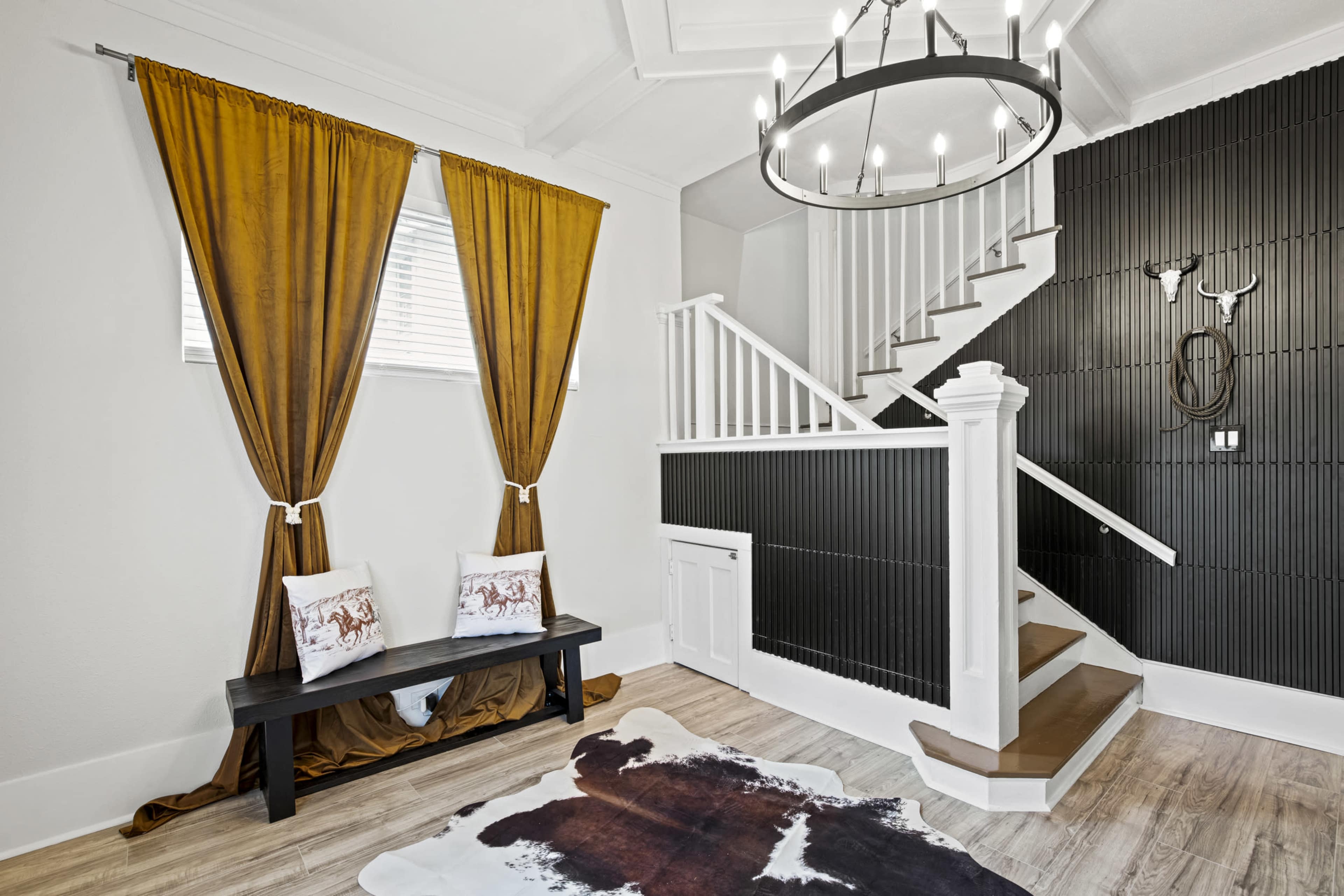 The image shows a bright entryway featuring a staircase, a black accent wall, a wooden bench with decorative pillows, and yellow curtains.