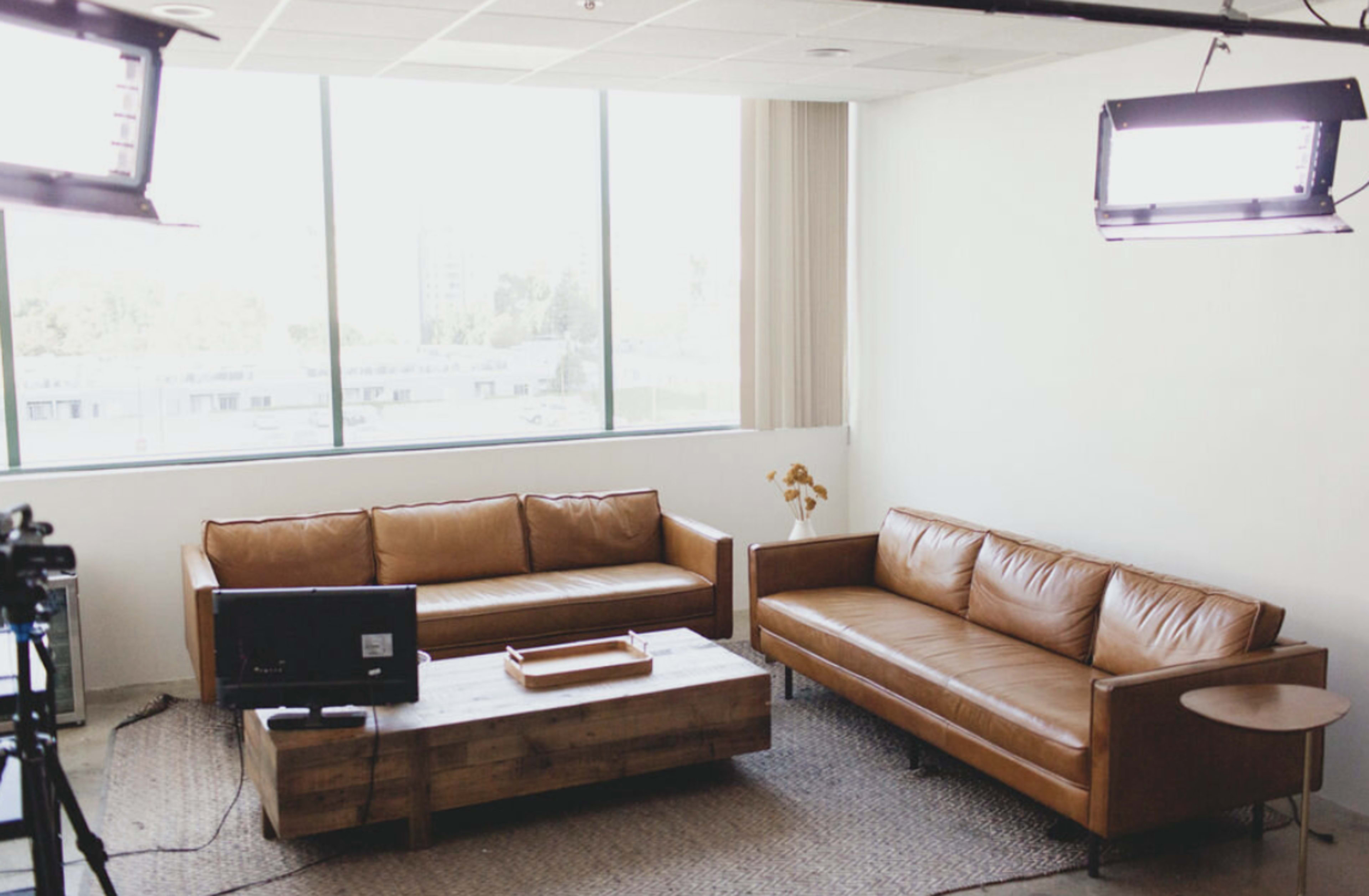 A bright, modern living room with two brown leather sofas, a wooden coffee table, and a large window letting in natural light.