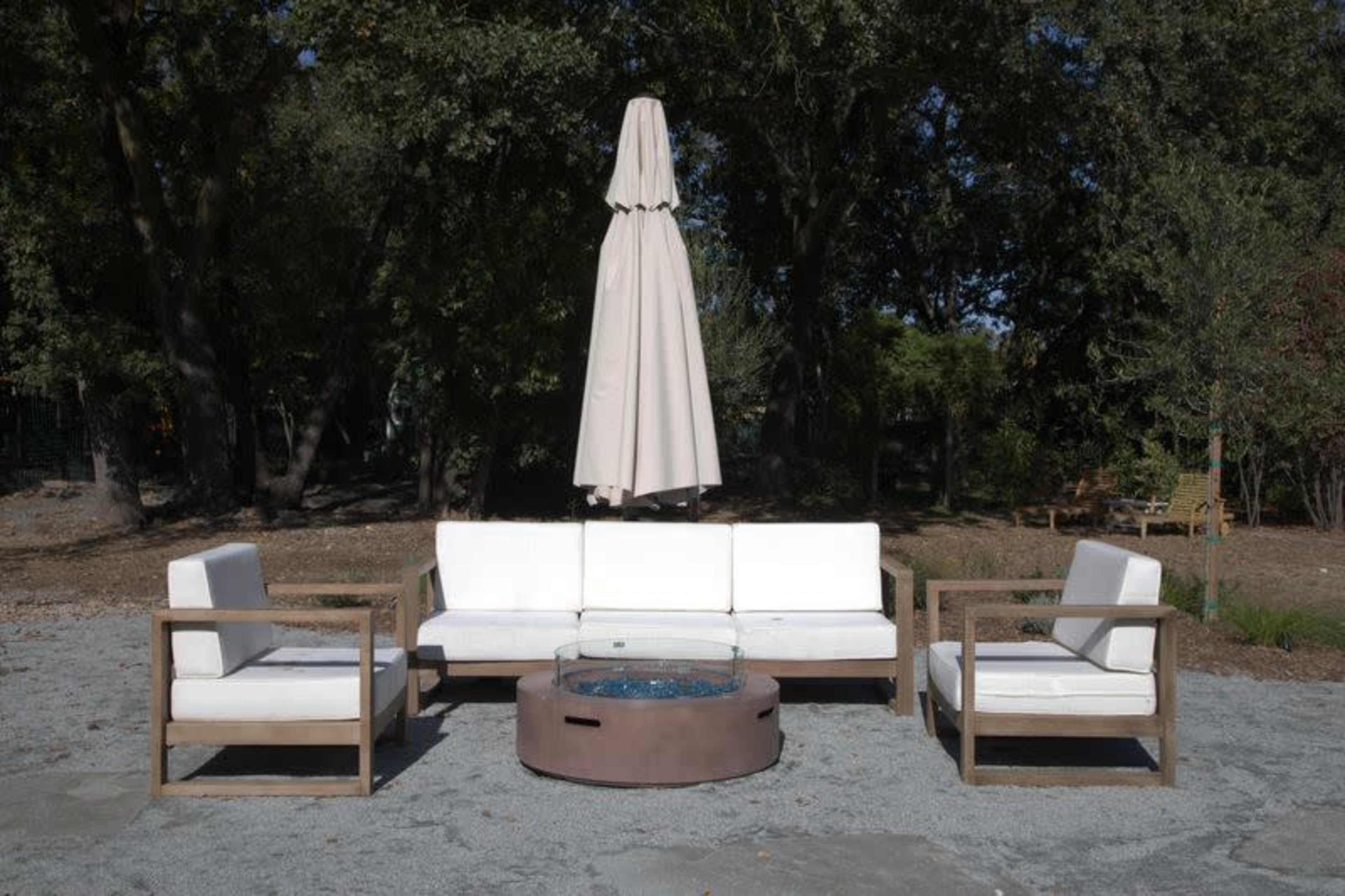 A modern outdoor seating arrangement features a sofa and two armchairs surrounding a circular fire pit, with a large umbrella positioned above.