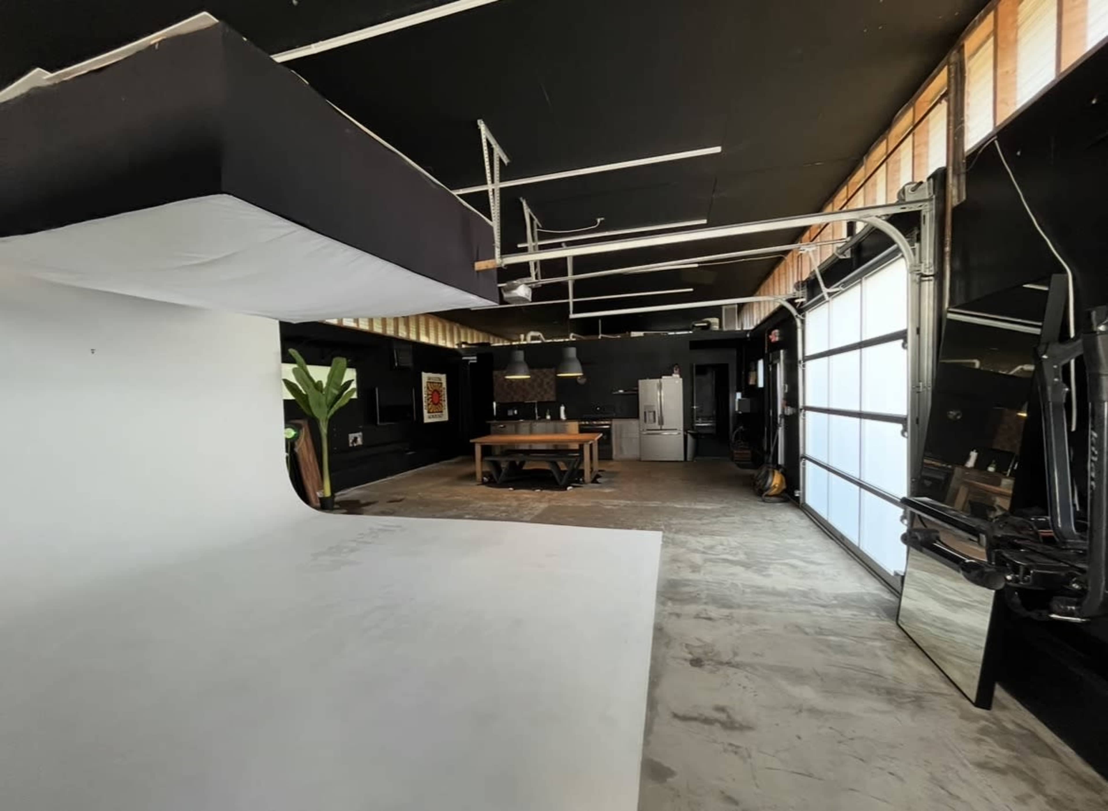 The image shows a spacious studio with a white backdrop on the left side, a large wooden table in the center, and a garage door leading to the outside on the right.