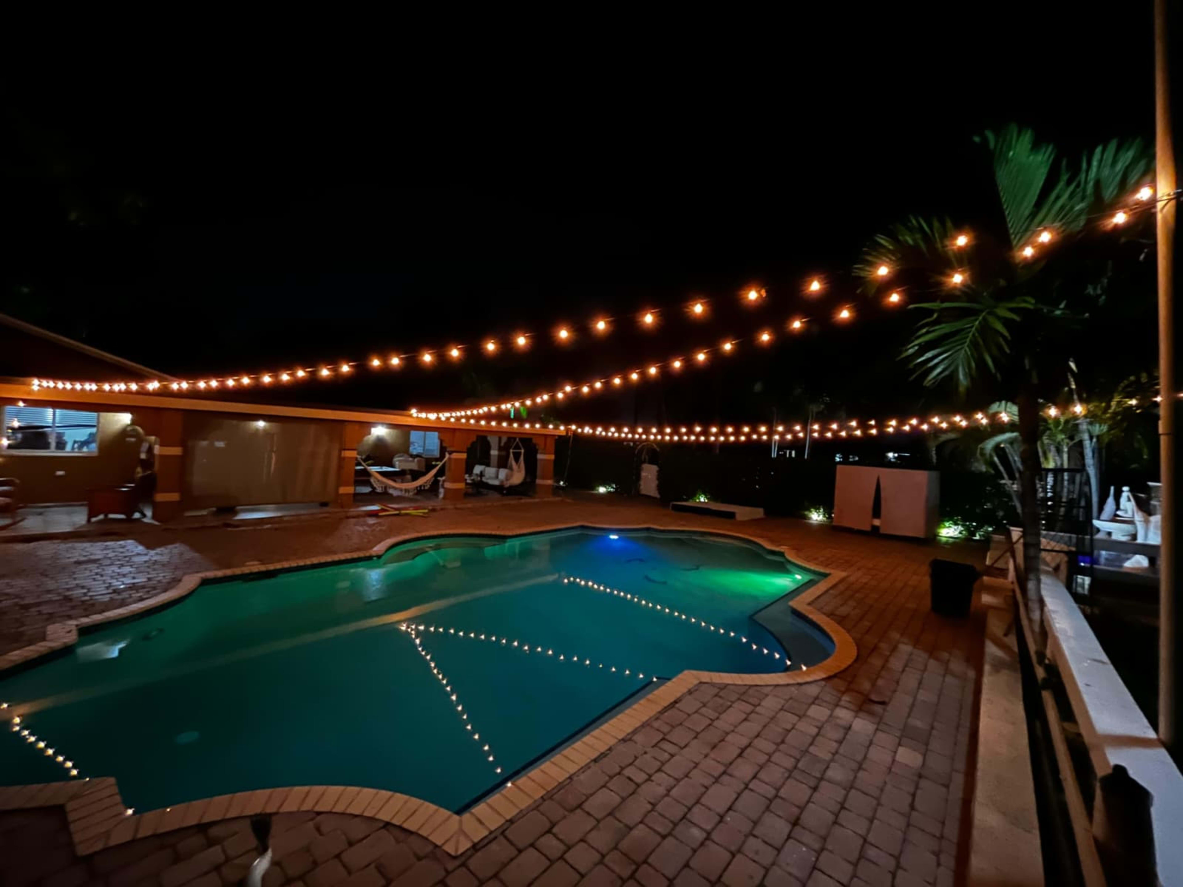 The image shows a well-lit pool area at night, featuring a neatly arranged pool surrounded by string lights and a patio.