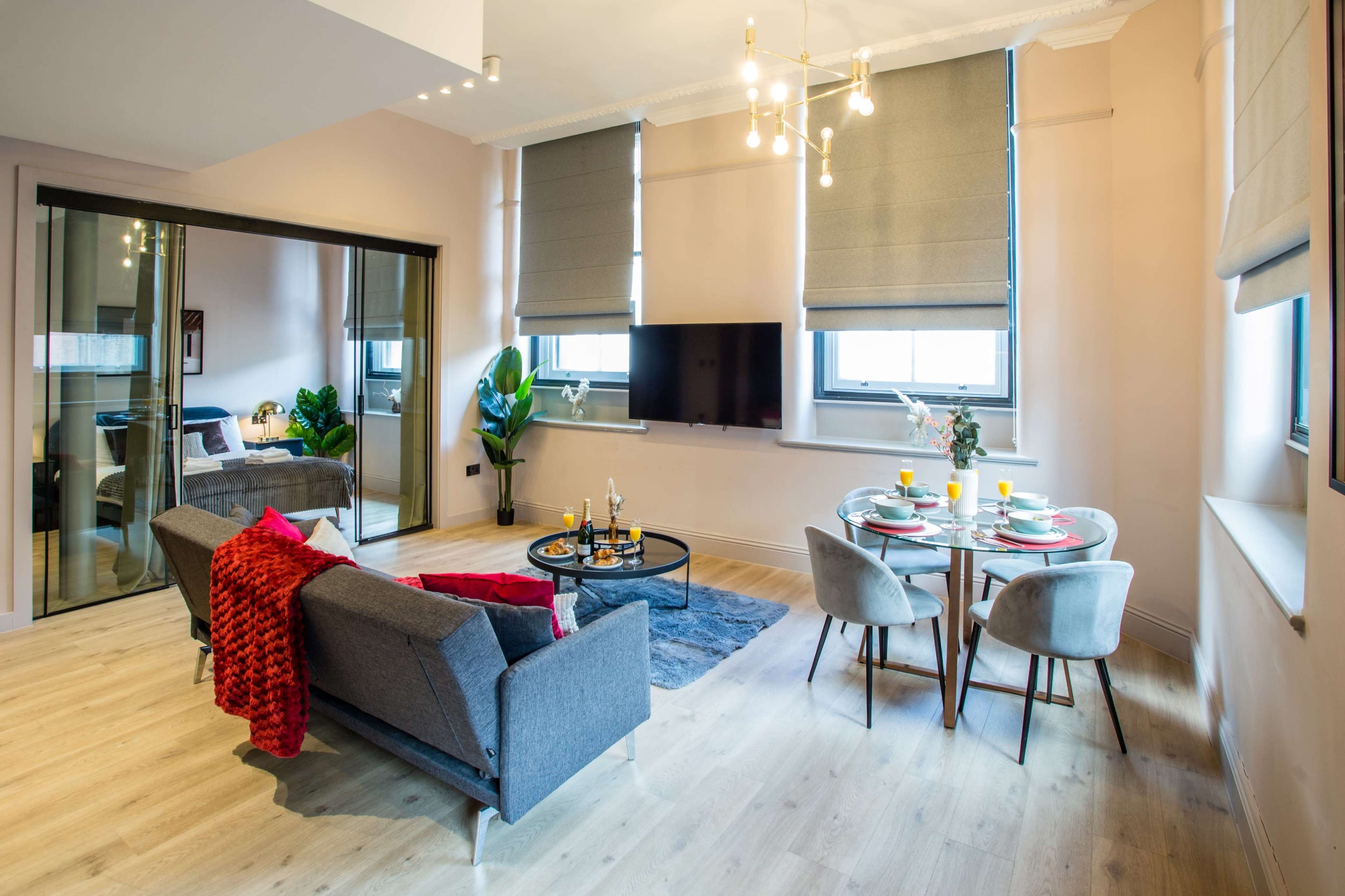 The image shows a modern living area featuring a gray sofa, a round dining table set for two, and large windows with light-filtering shades.