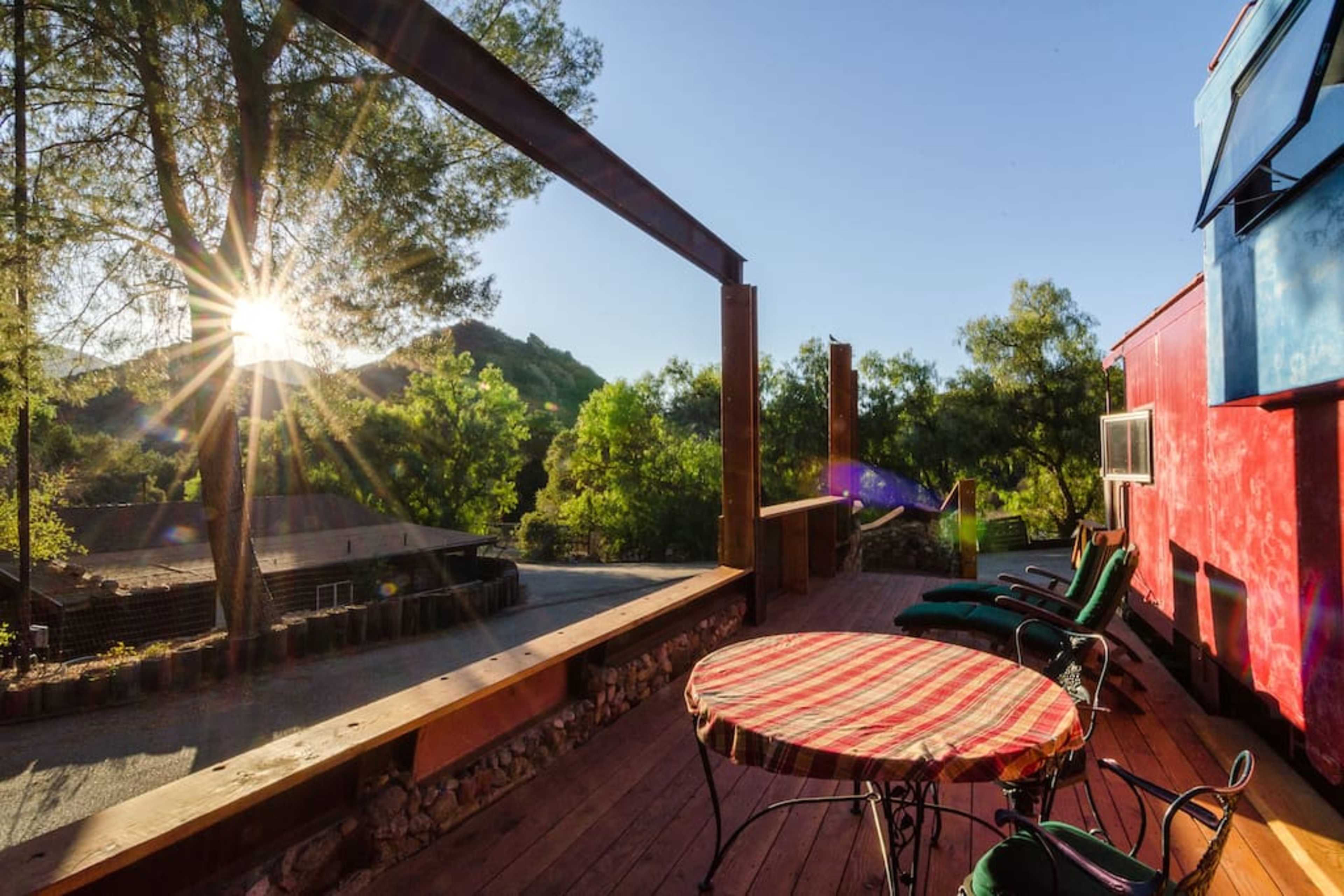 A deck with a round table and lounge chairs overlooks a landscape with trees and mountains, illuminated by sunlight.