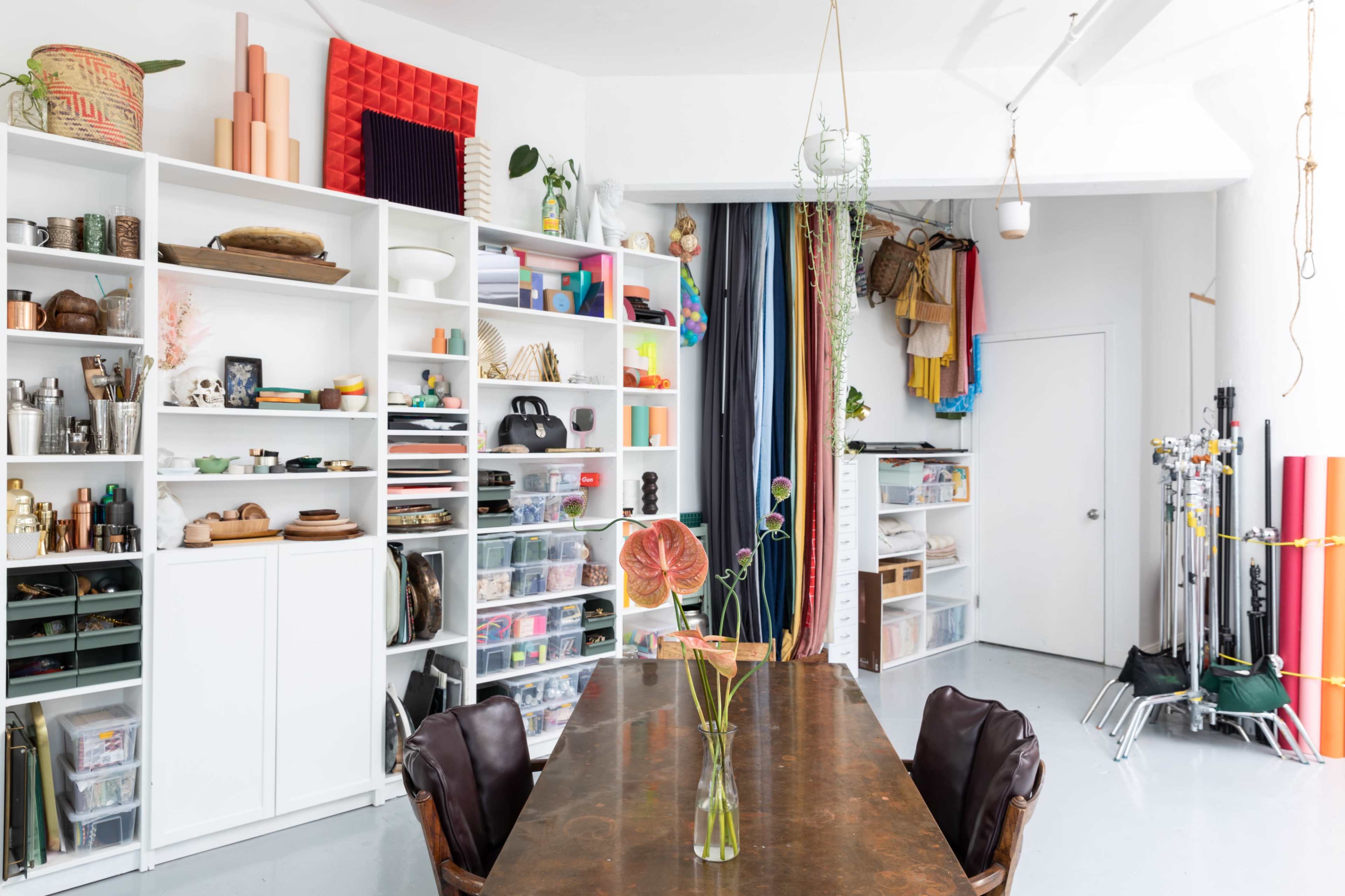 A bright, organized studio space with a large wooden table at the center, surrounded by shelves filled with various craft supplies and decorative items.