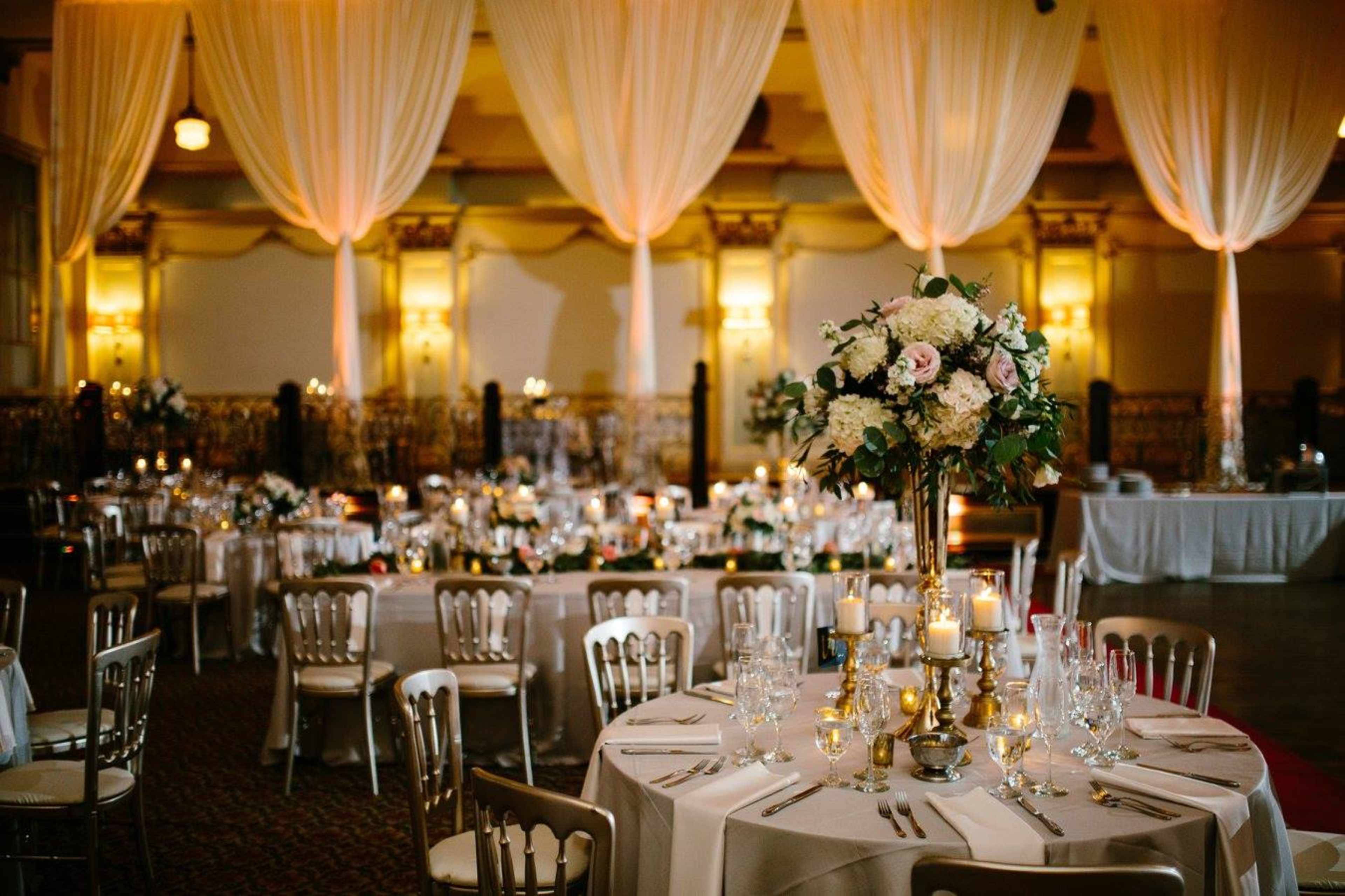 A banquet hall is set for an event, featuring elegantly arranged tables with floral centerpieces, candles, and draped fabric overhead.