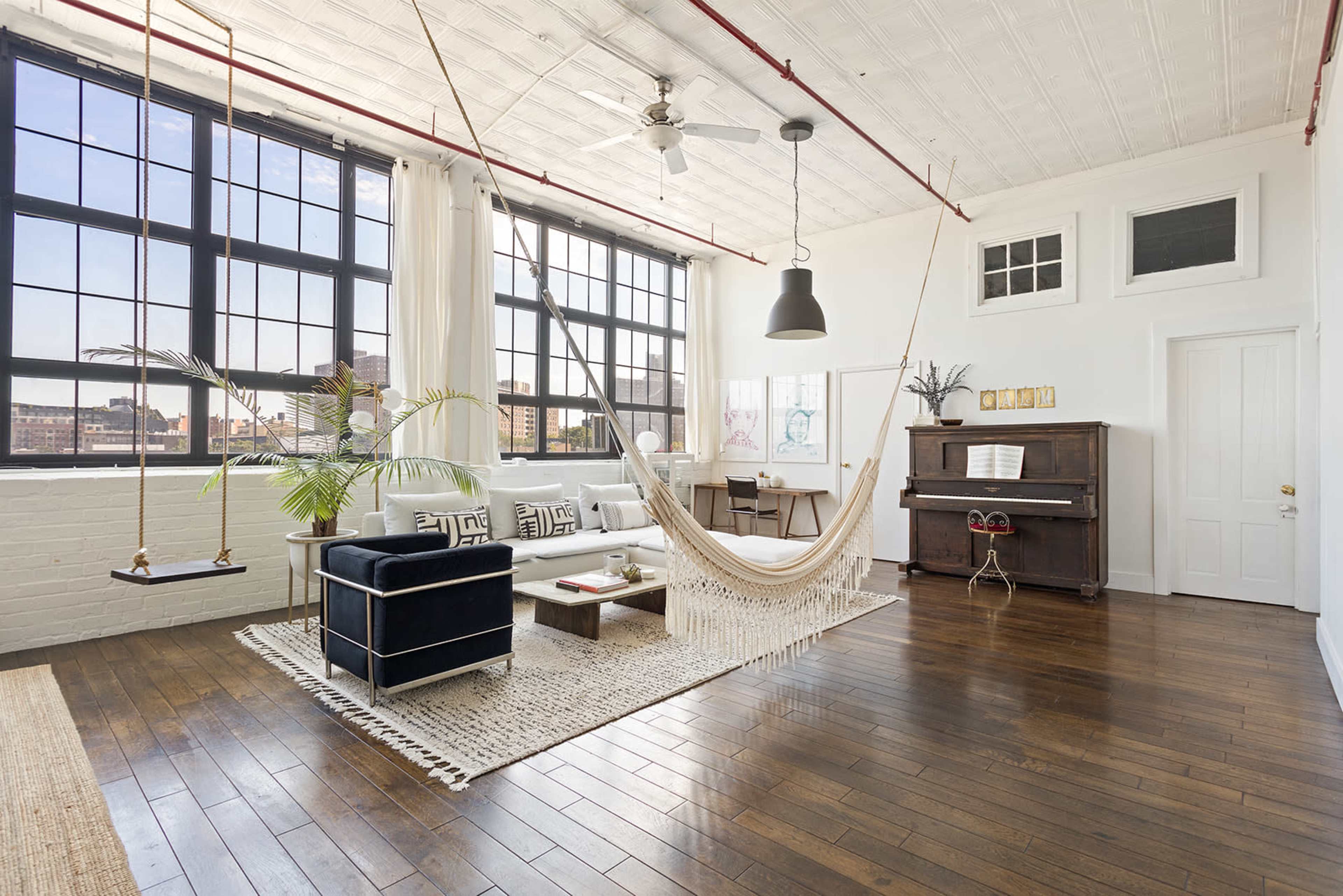 A spacious, well-lit living area featuring a hammock suspended from the ceiling, a dark blue armchair, a wooden piano, and large windows overlooking a cityscape.
