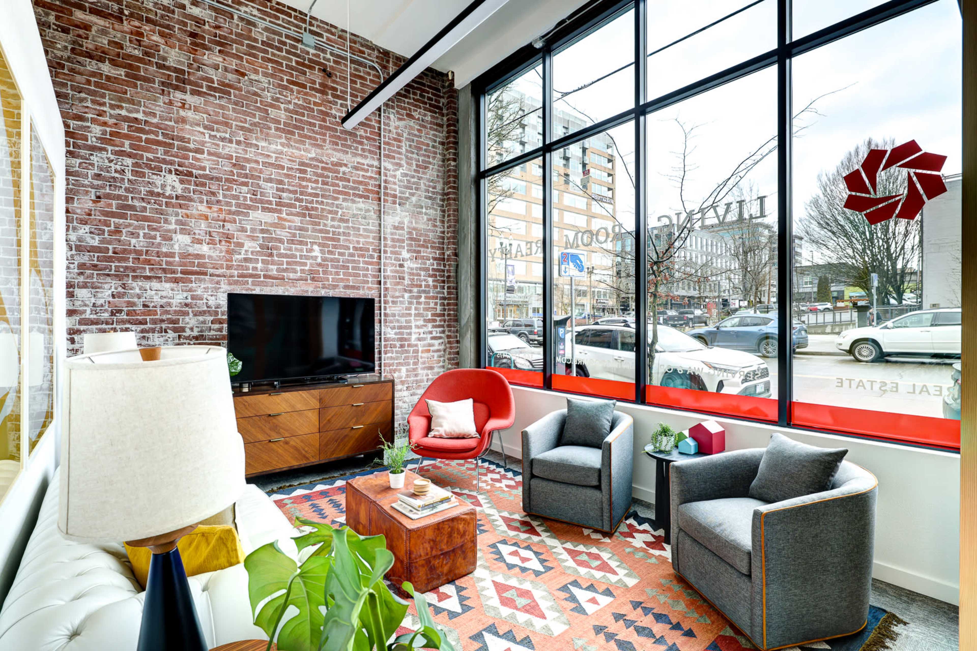 A modern lounge area with a brick wall, large windows, a television, two armchairs, a coffee table, and decorative plants.