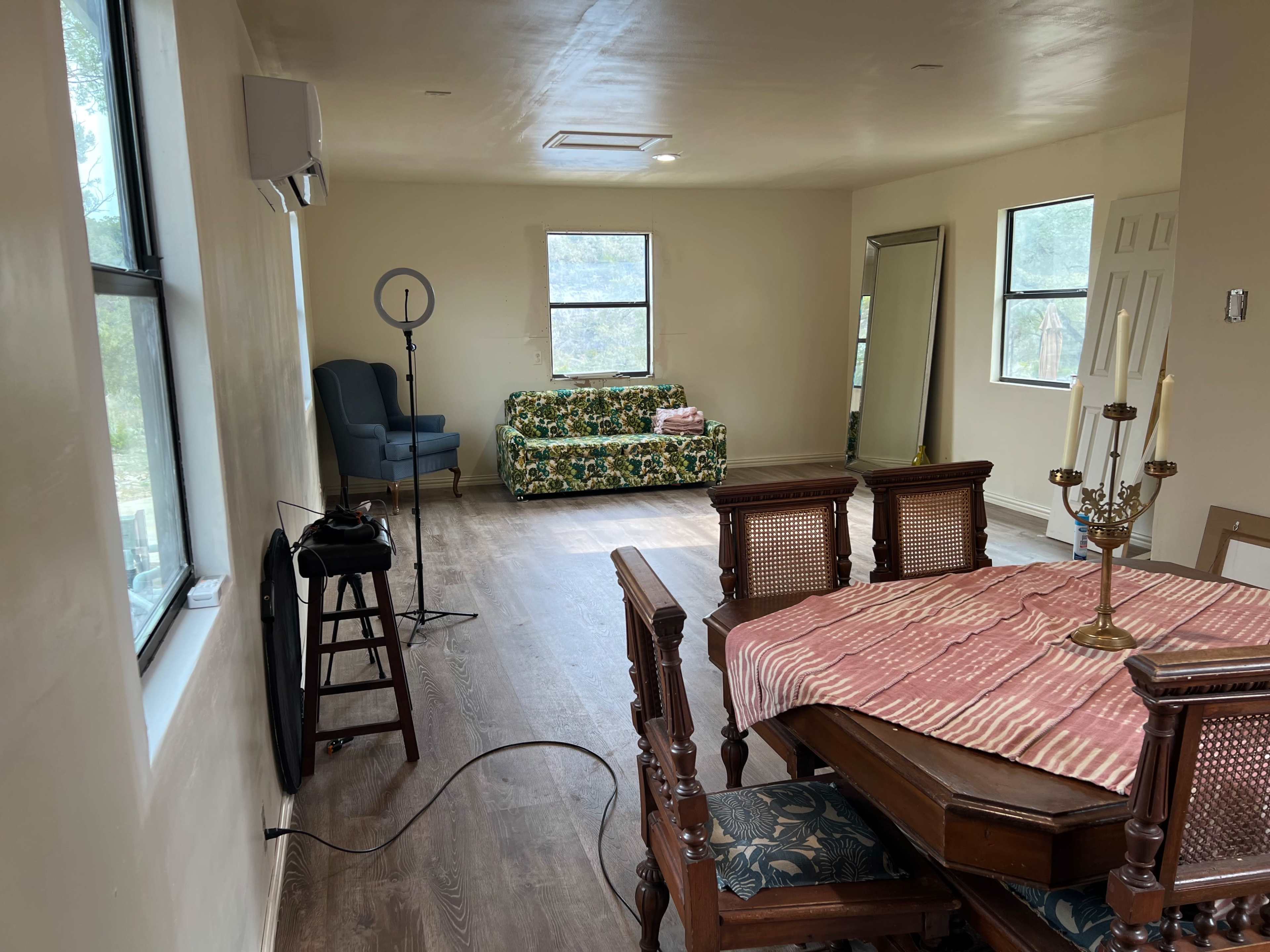 A spacious room features a dining table with chairs, a floral couch, a mirror, and a standing lamp.