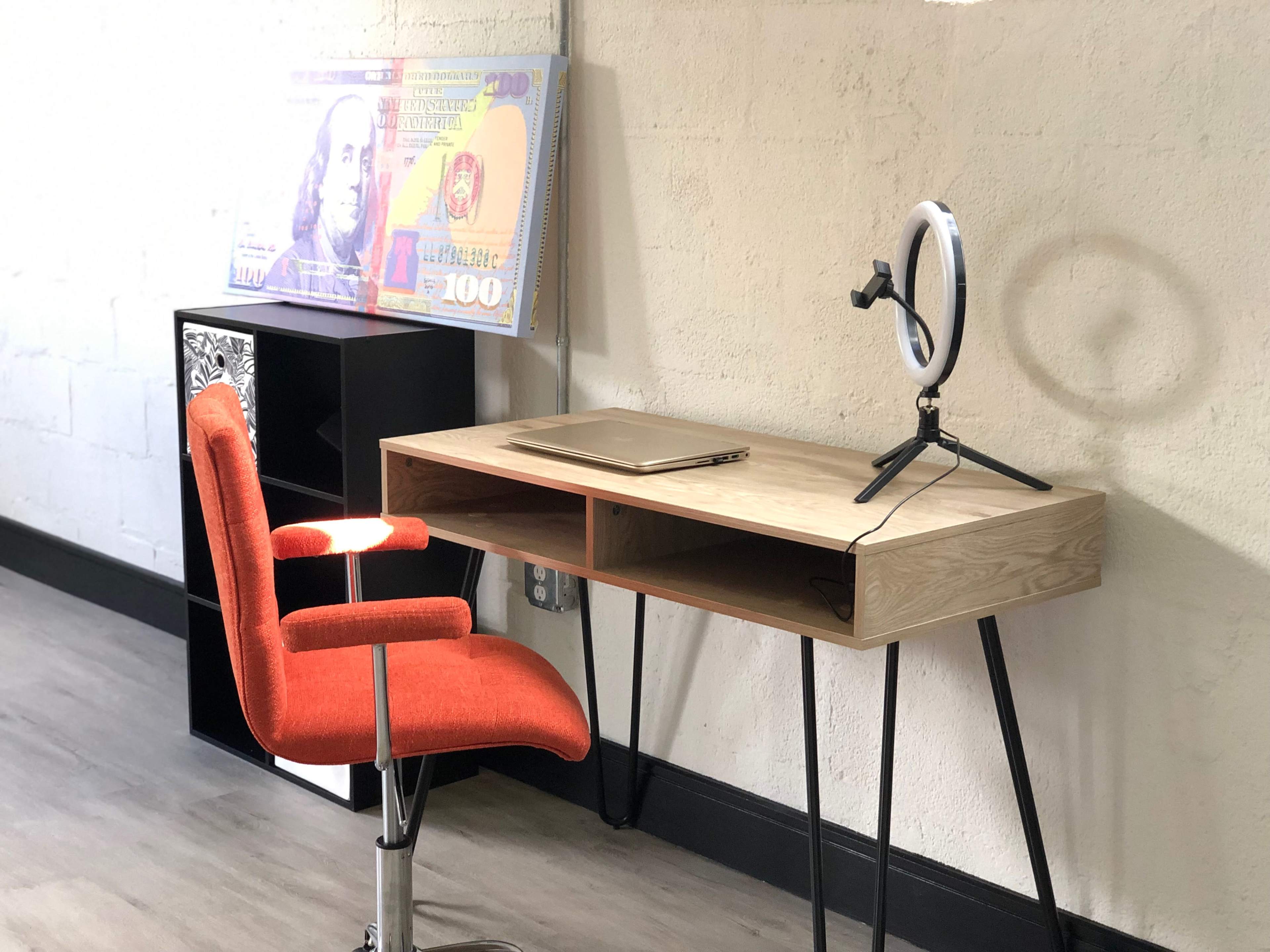 A modern wooden desk with an orange chair and a ring light stands against a white wall, alongside a large decorative banknote.