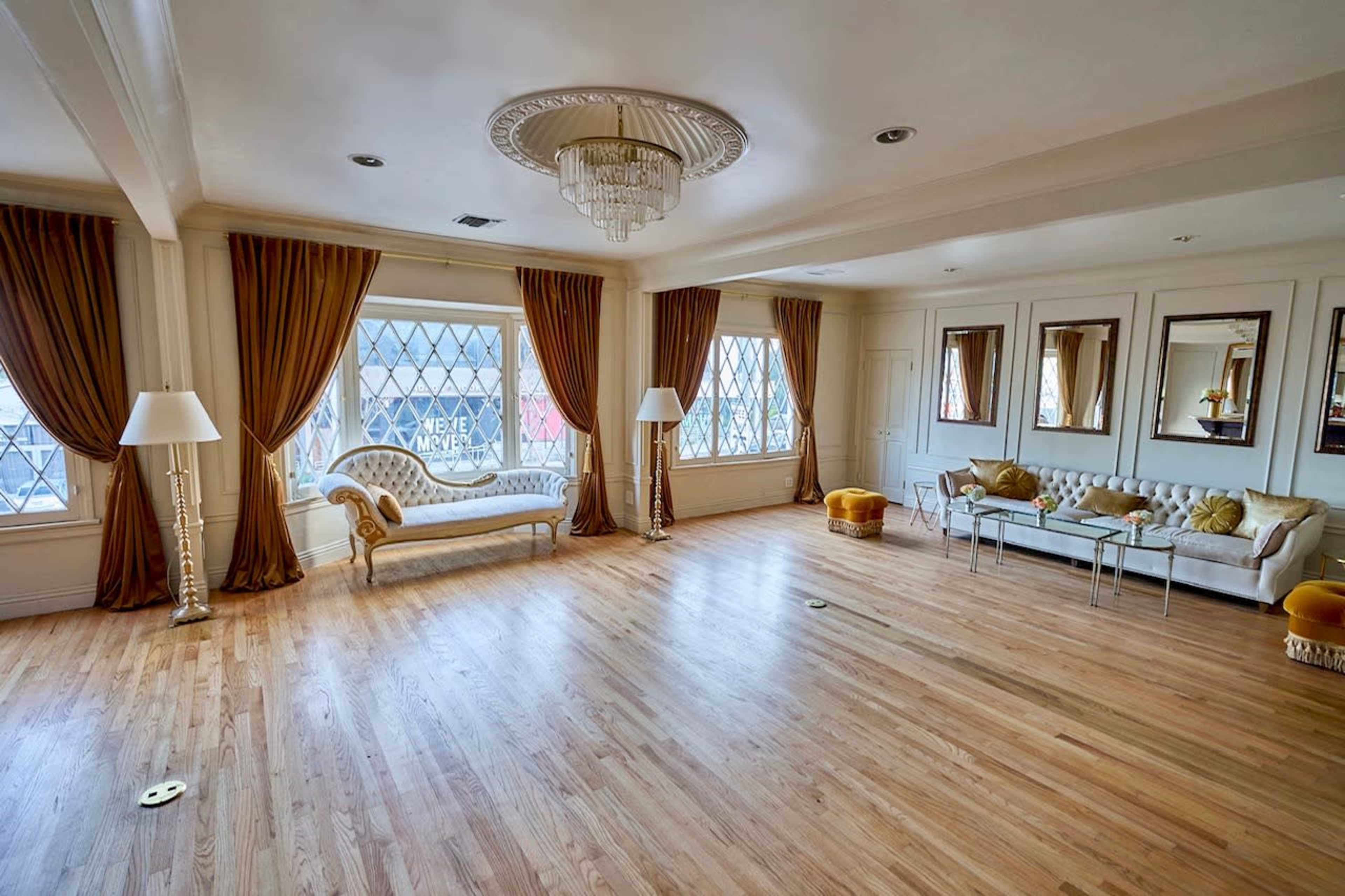 The image shows a spacious, well-lit room with large windows, hardwood flooring, decorative mirrors, and elegant furniture.