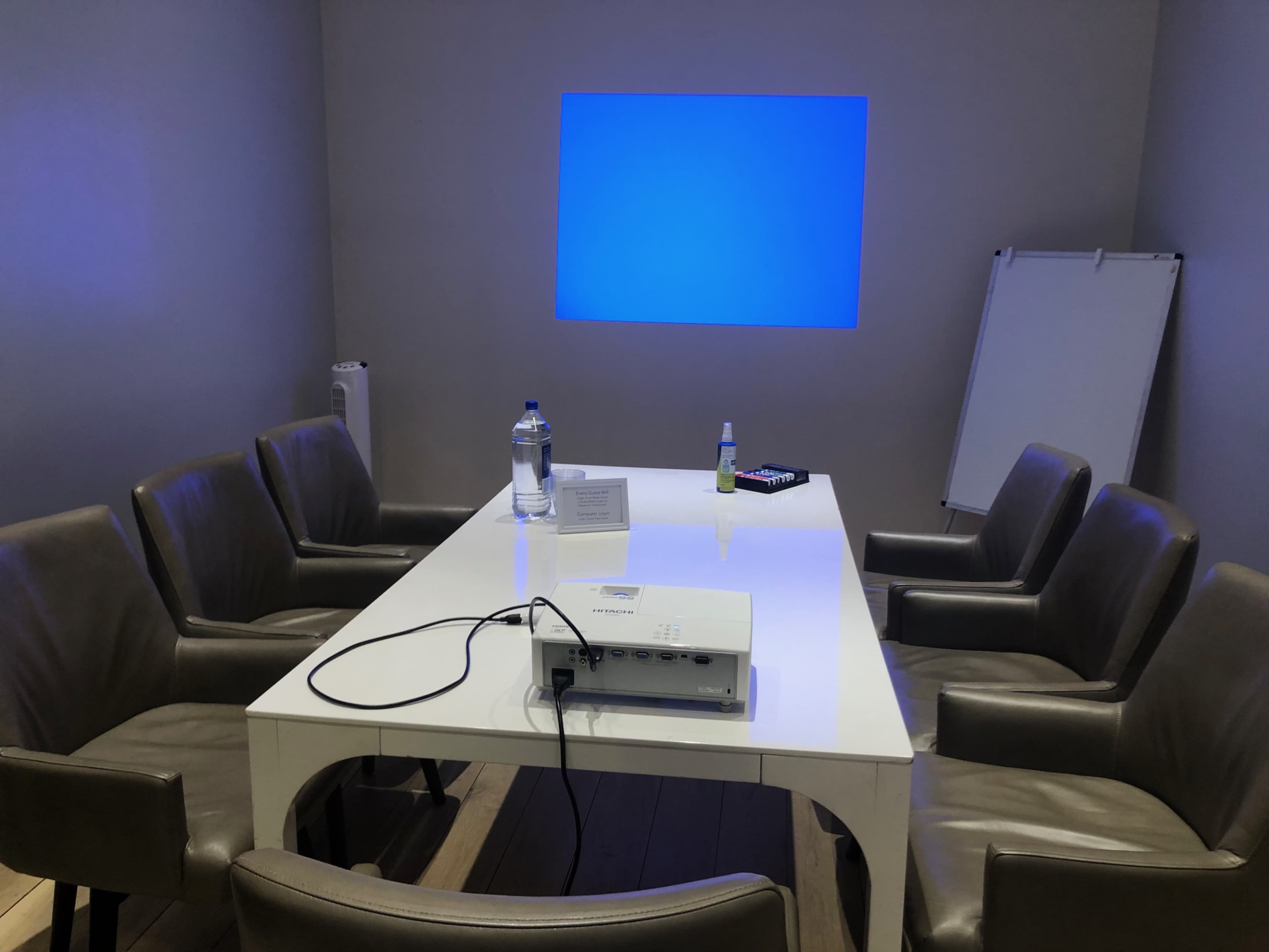 A conference room features a rectangular white table surrounded by six brown chairs, with a projection screen displaying a blue background.