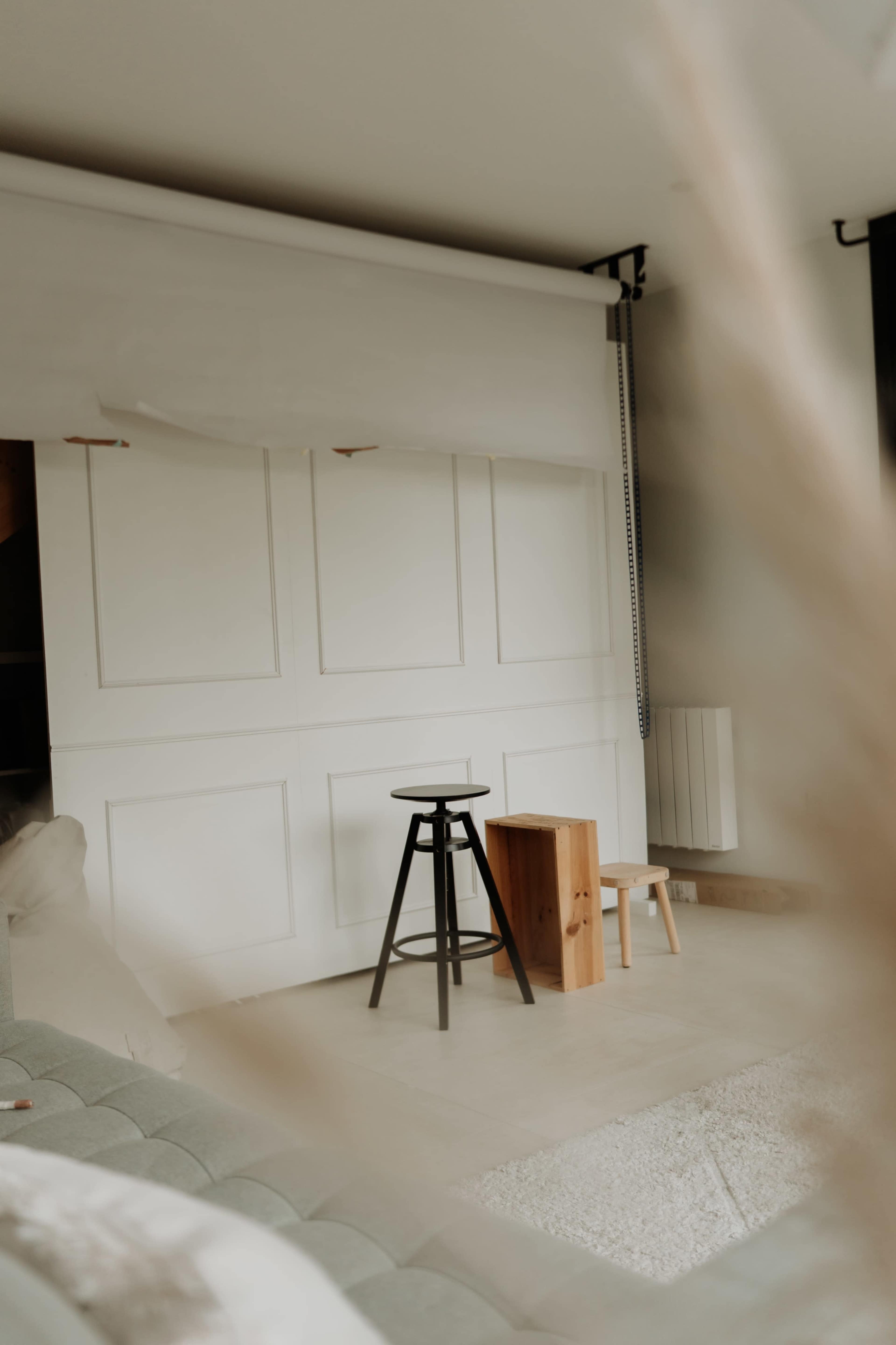 The image shows a minimalistic indoor setting with a black stool and two wooden blocks, set against a white panel wall and a slightly blurred foreground.