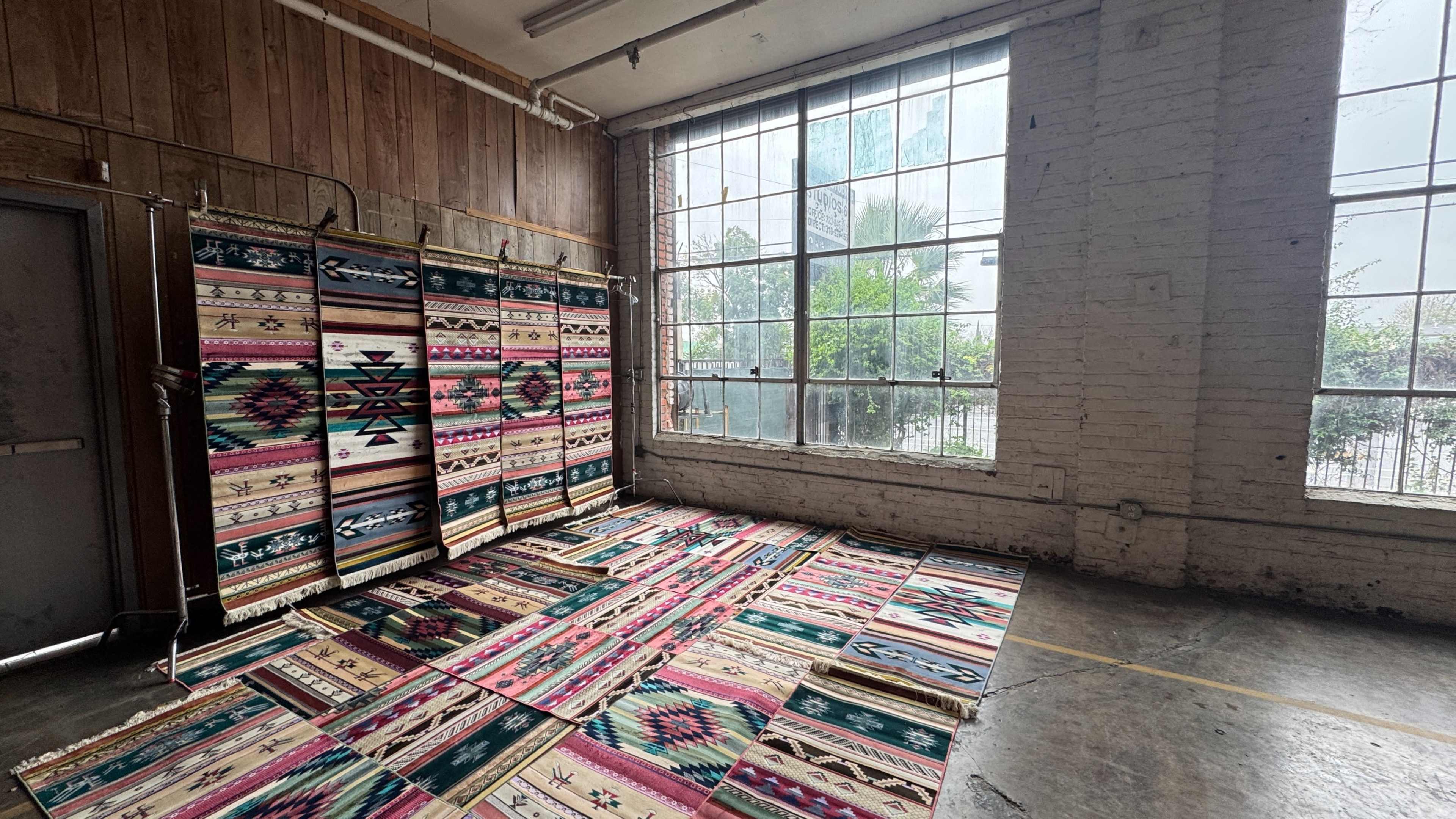 The image shows a room with large windows, featuring colorful woven rugs arranged on the floor and hanging on the walls.