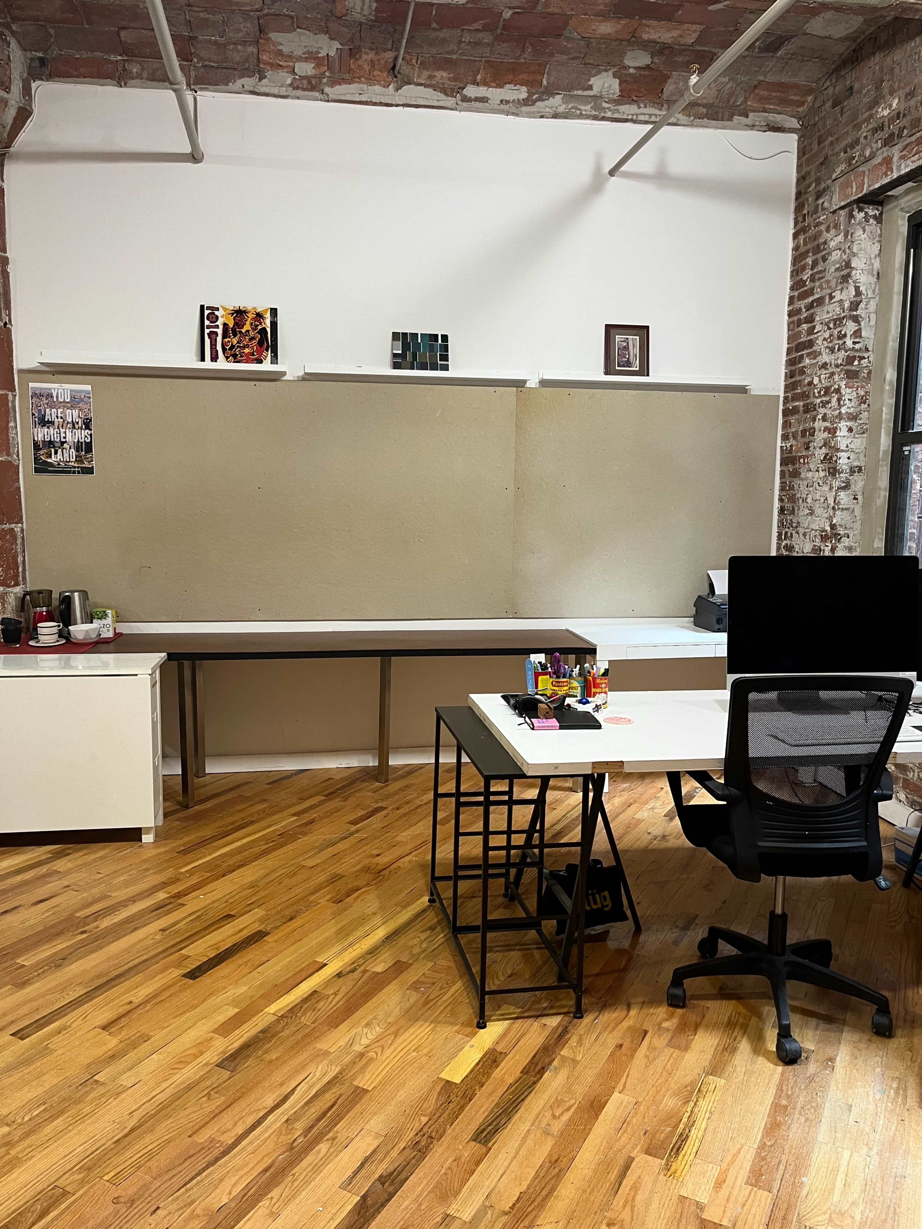 A modern office space featuring a desk with a computer, a chair, and a long table against a wall adorned with a few framed pictures.