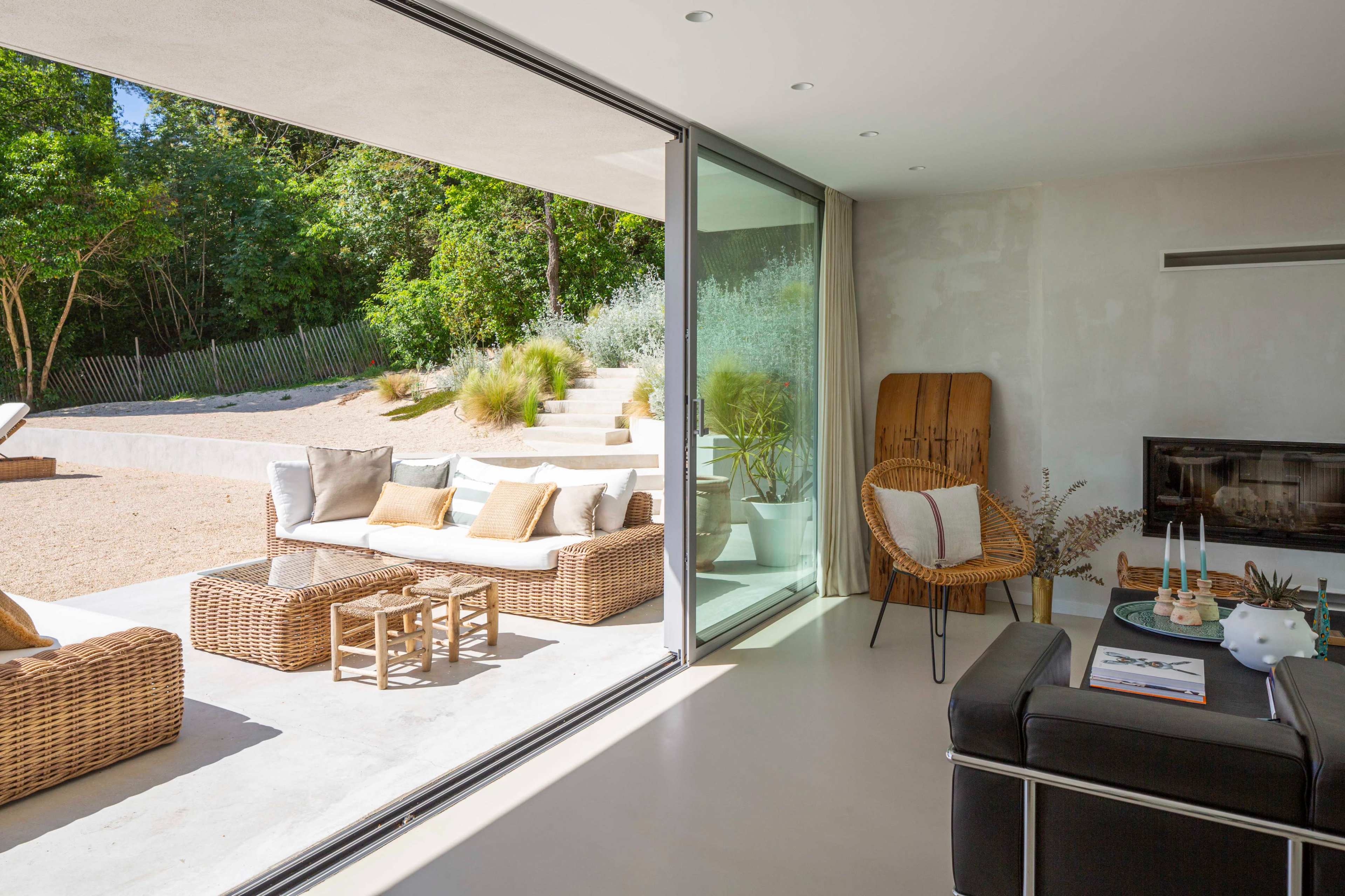 The image shows a modern living space with large glass windows offering a view of an outdoor seating area surrounded by greenery.
