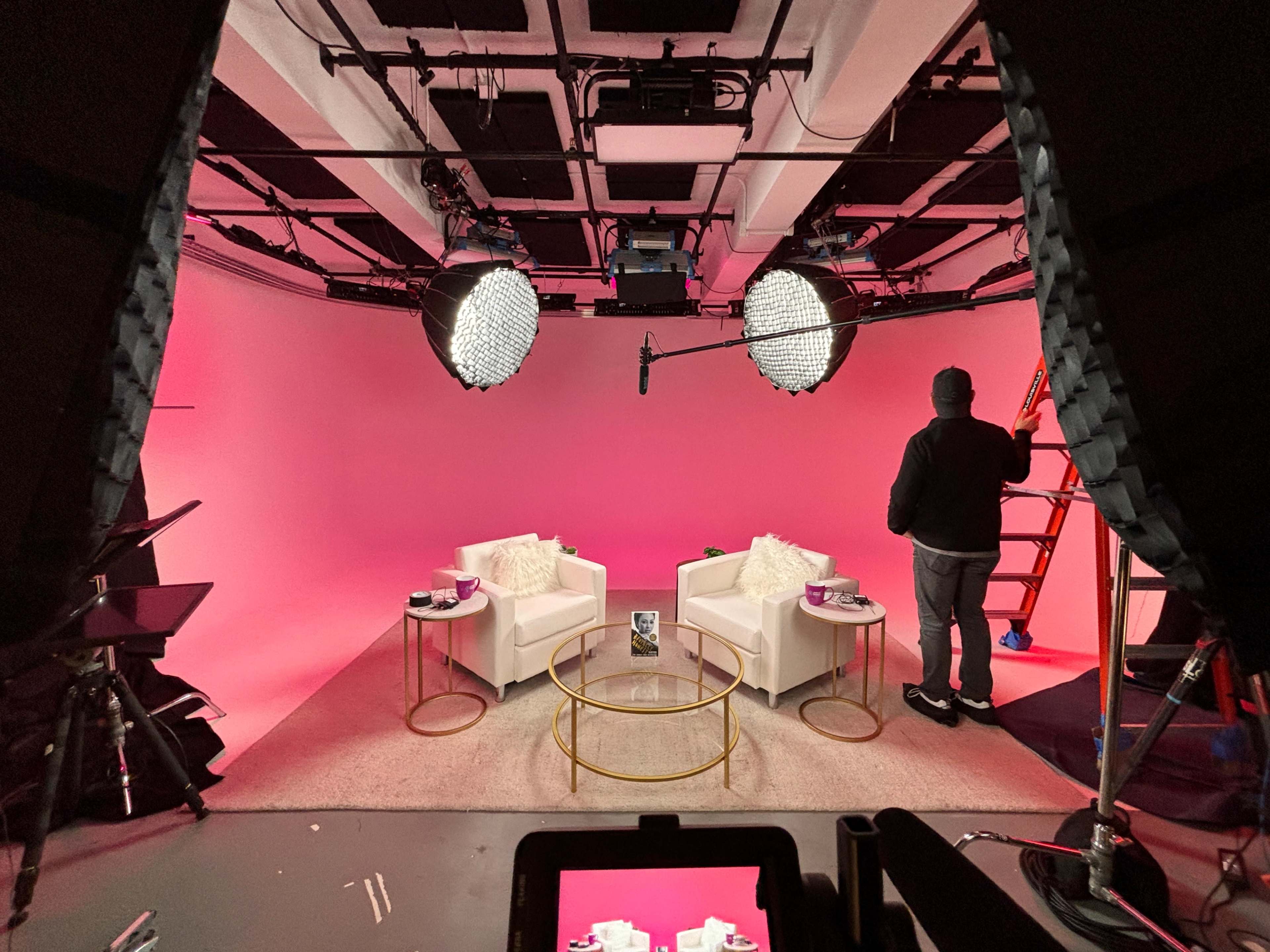 A television studio set features two white chairs with decorative pillows, a glass coffee table, and bright pink background lighting, while a person adjusts equipment on a ladder.